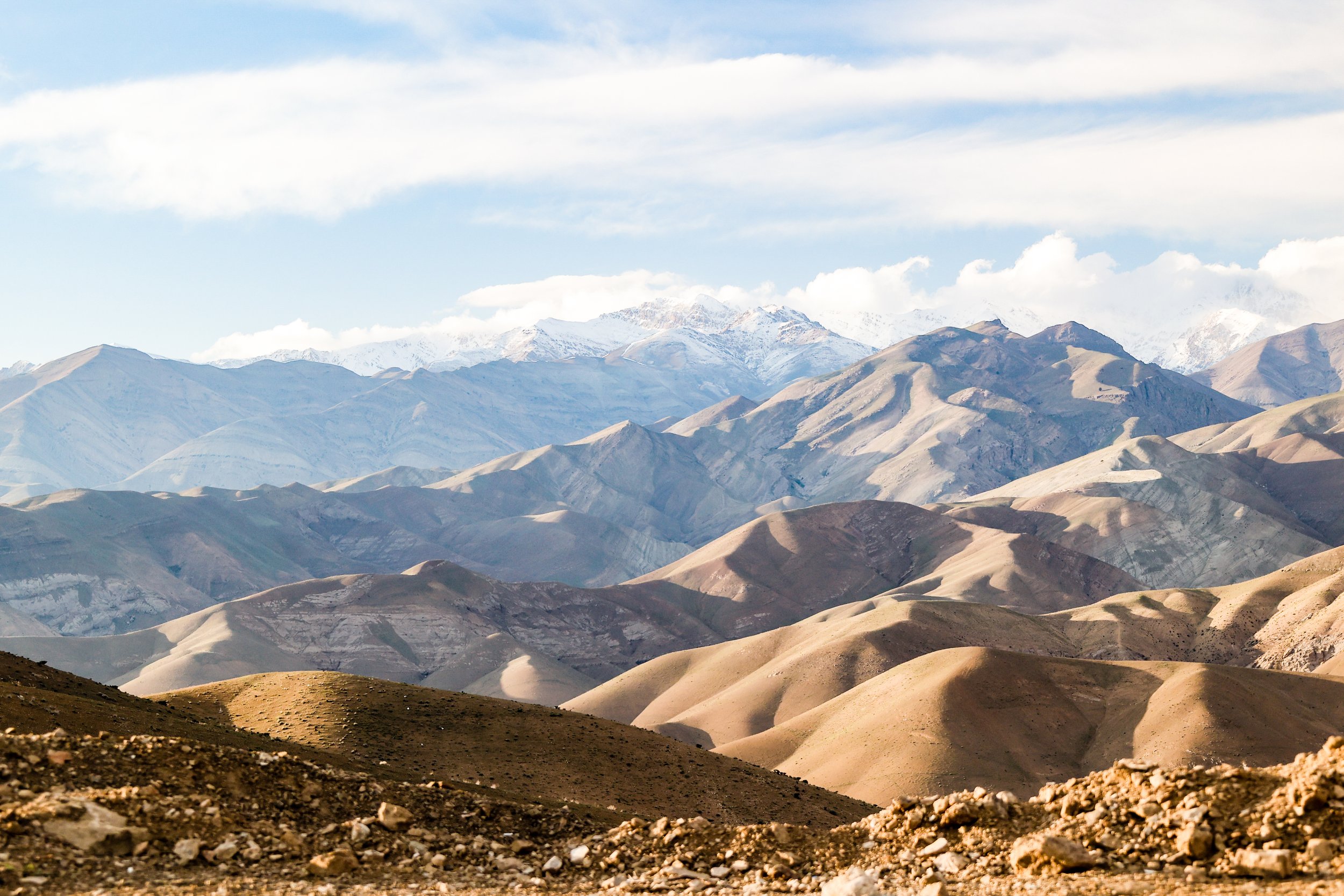 Scenic view of layered desert mountains with snow-capped peaks in the background under a partly cloudy sky.