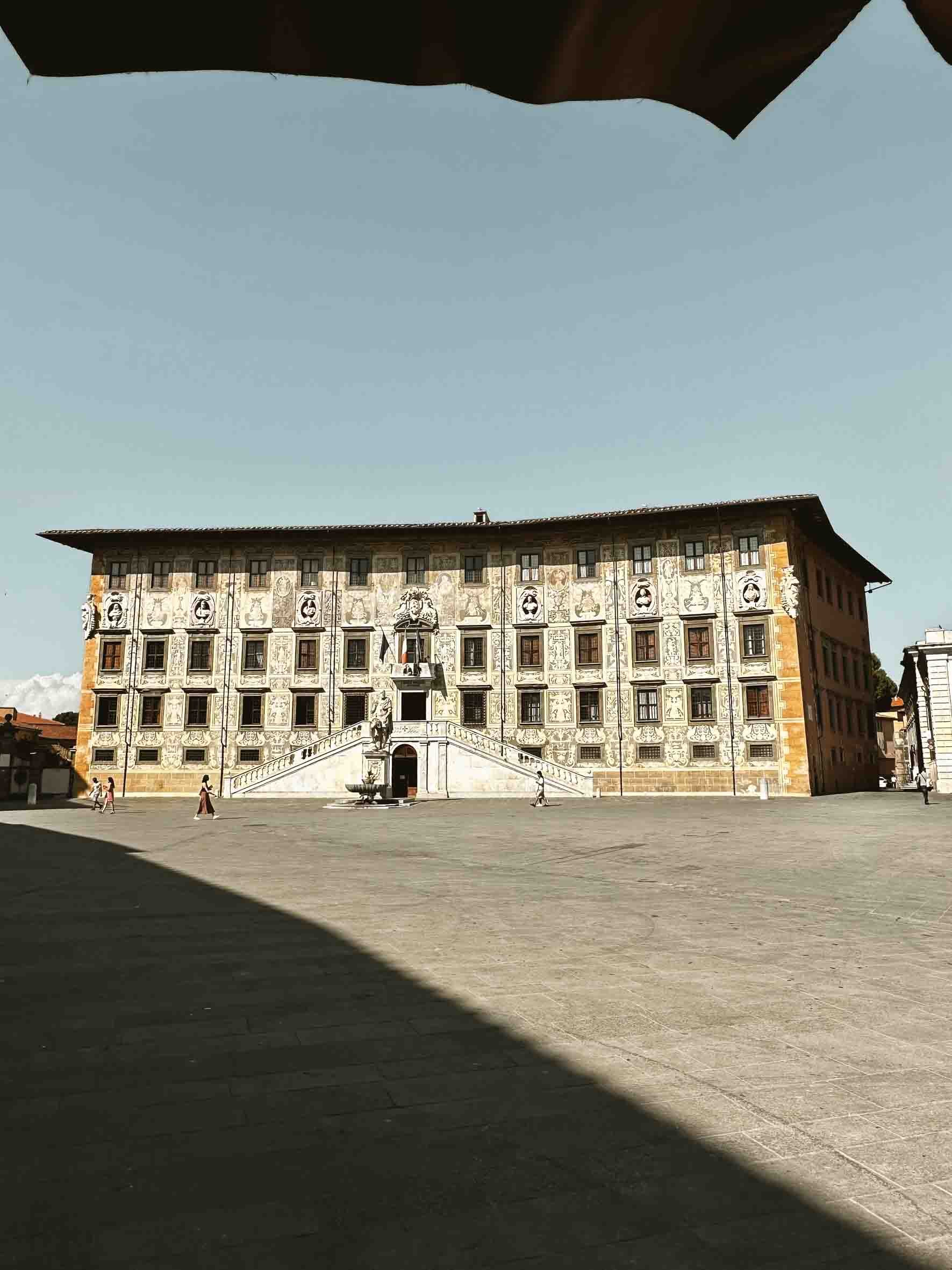 A historic building with ornate facade, staircase entrance, and sculptures in a European square, with shadows on the ground and a clear sky.
