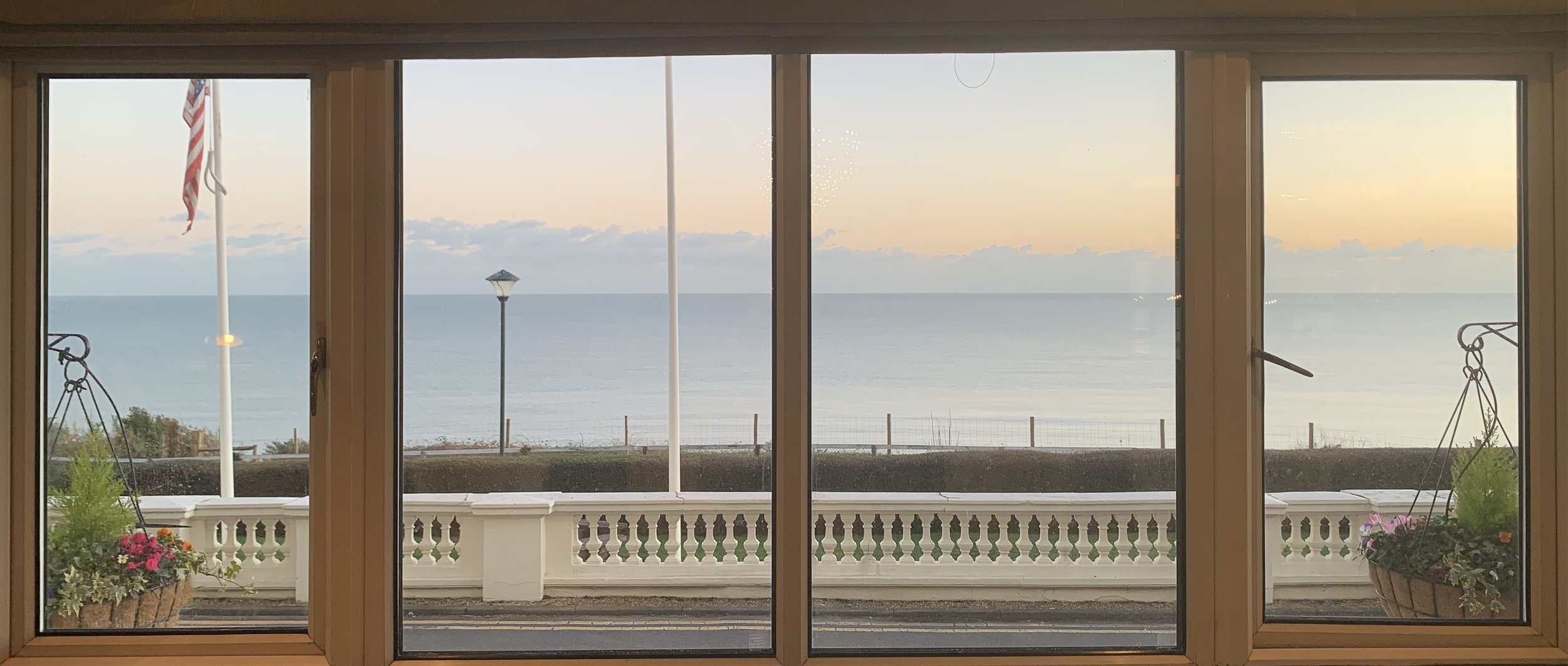 View through a window of a balcony overlooking the ocean at sunset, with potted flowers, a white railing, and flagpole with an American flag.