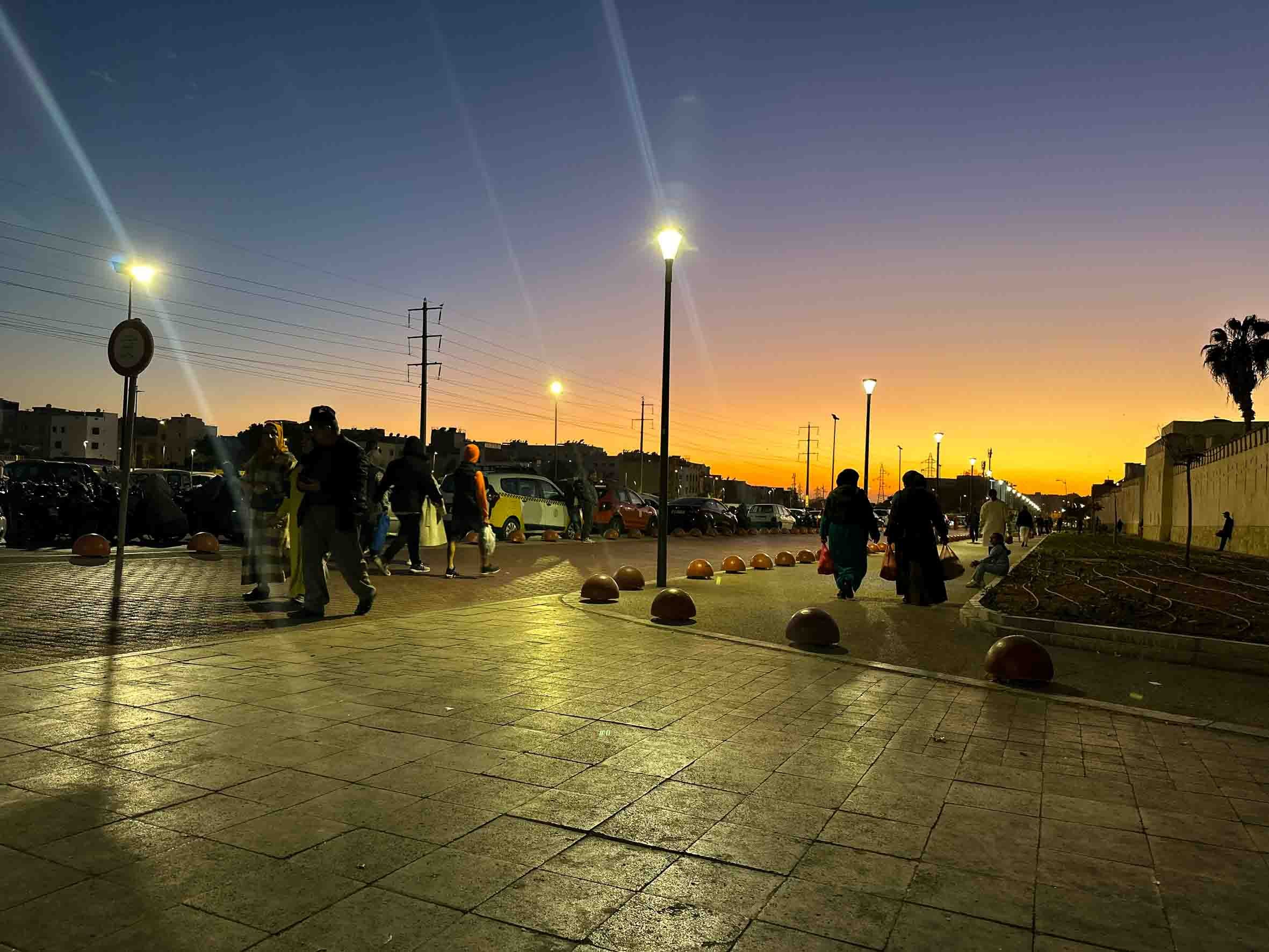 People walking outside at sunset with streetlights on and parked cars nearby