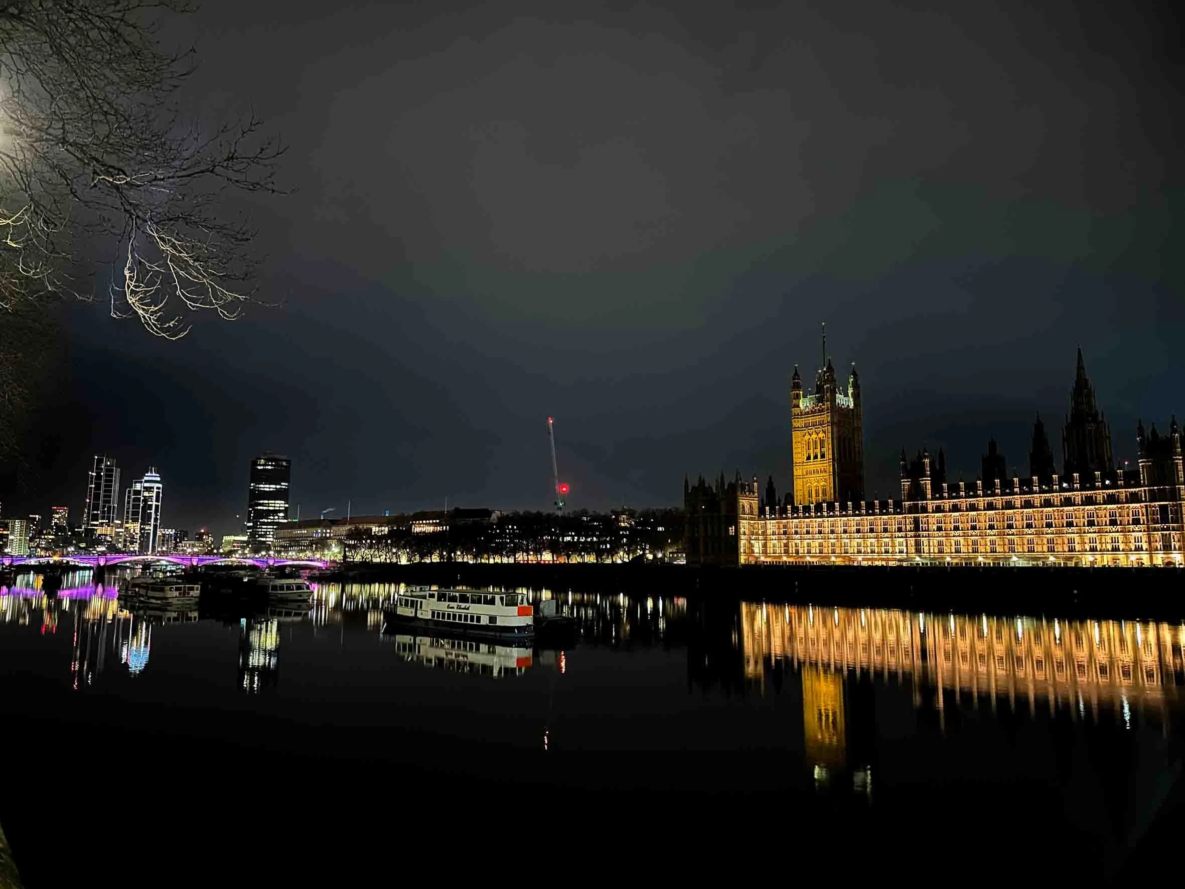 Nighttime view of London skyline with illuminated buildings, the Houses of Parliament, and reflections on the River Thames.