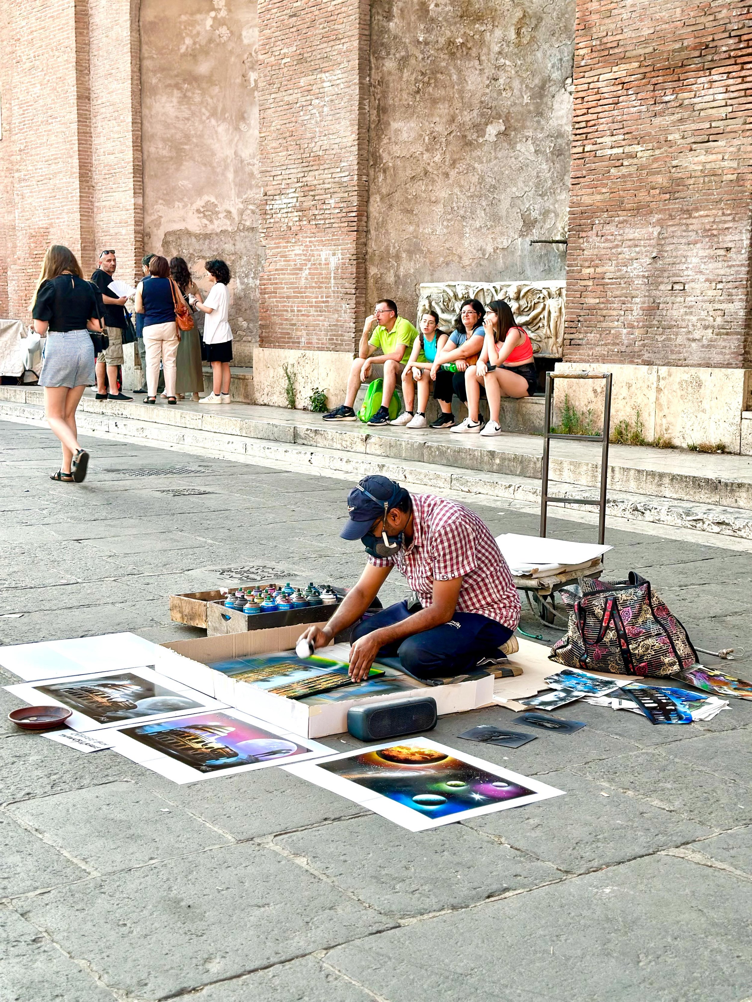 Street artist creating graffiti art on the pavement with spray paint, surrounded by colorful prints, spectators sitting on the ledge, and people walking in the background near an old brick wall.