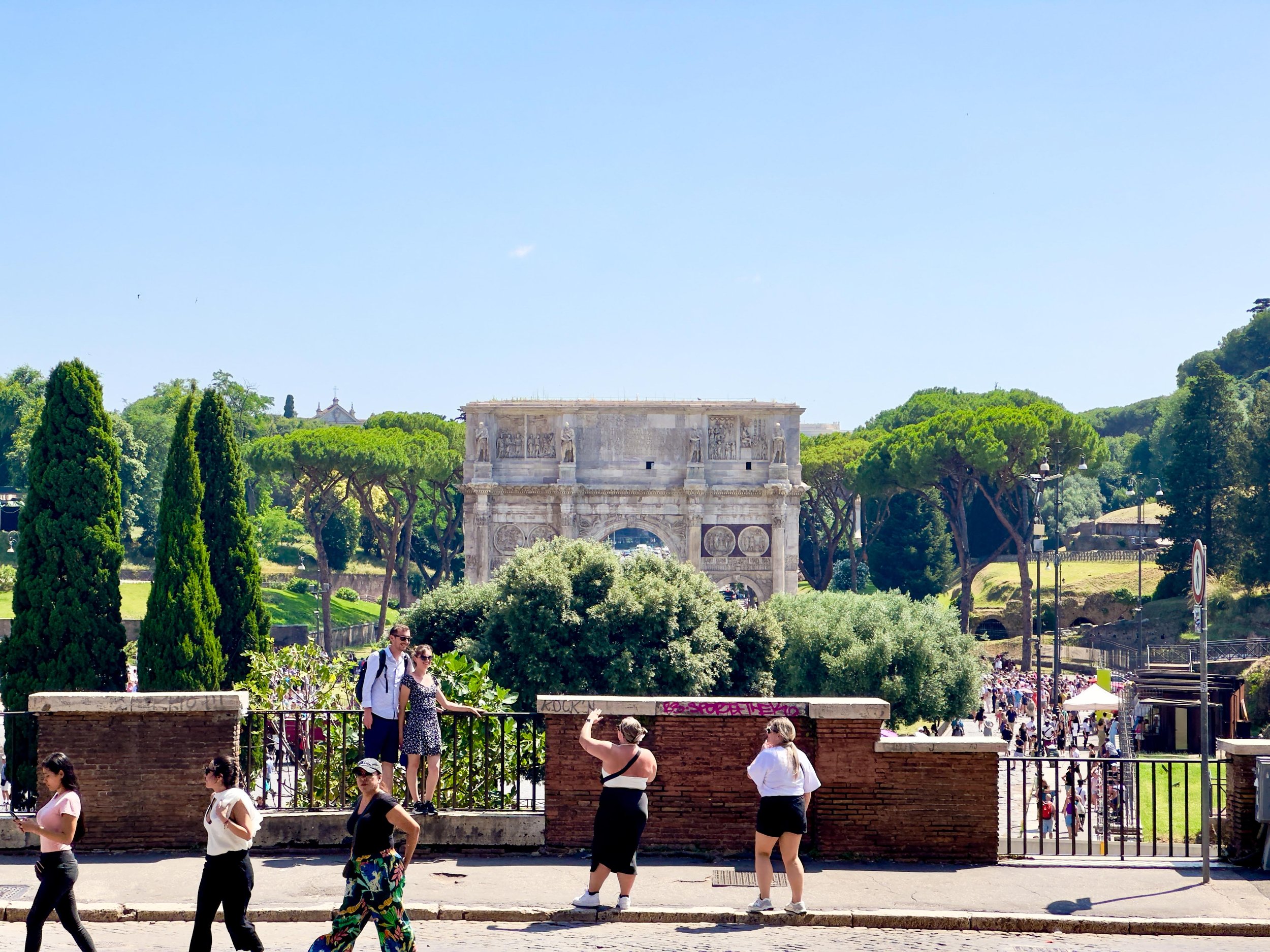 People walking and taking photos in front of the Arco di Constantino with trees and the Roman Forum in Rome, Italy, under a clear blue sky.