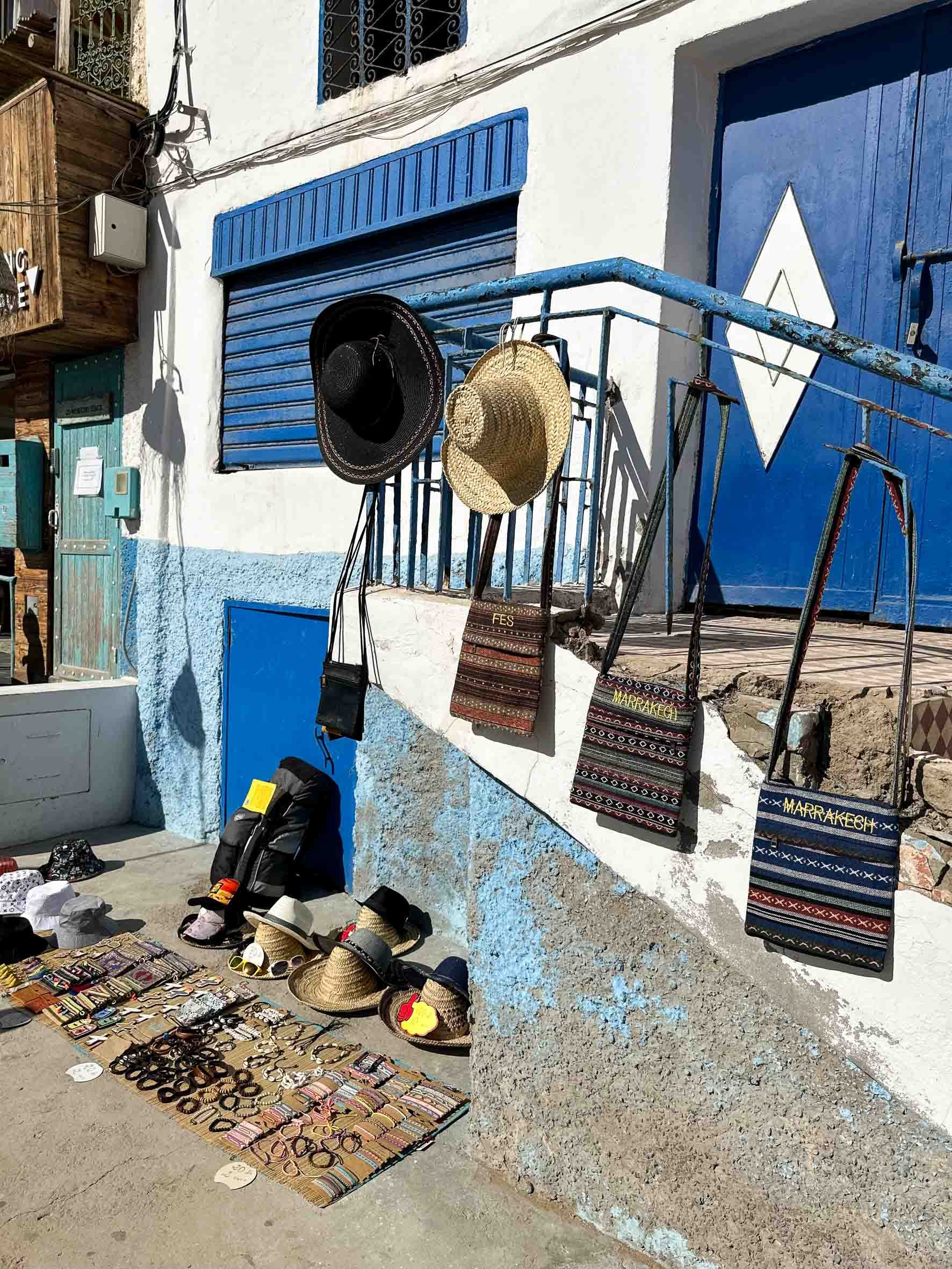 Street vendor selling hats, bags, jewelry, and textiles outside a building with blue window shutters and a blue door, in a sunny setting.