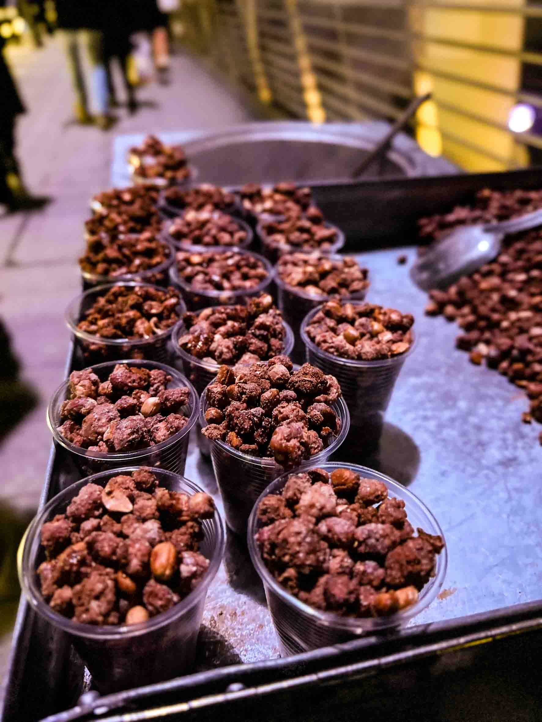 Multiple small plastic cups filled with trail mix containing nuts and dried fruit on a metal surface, with a blurred outdoor background.