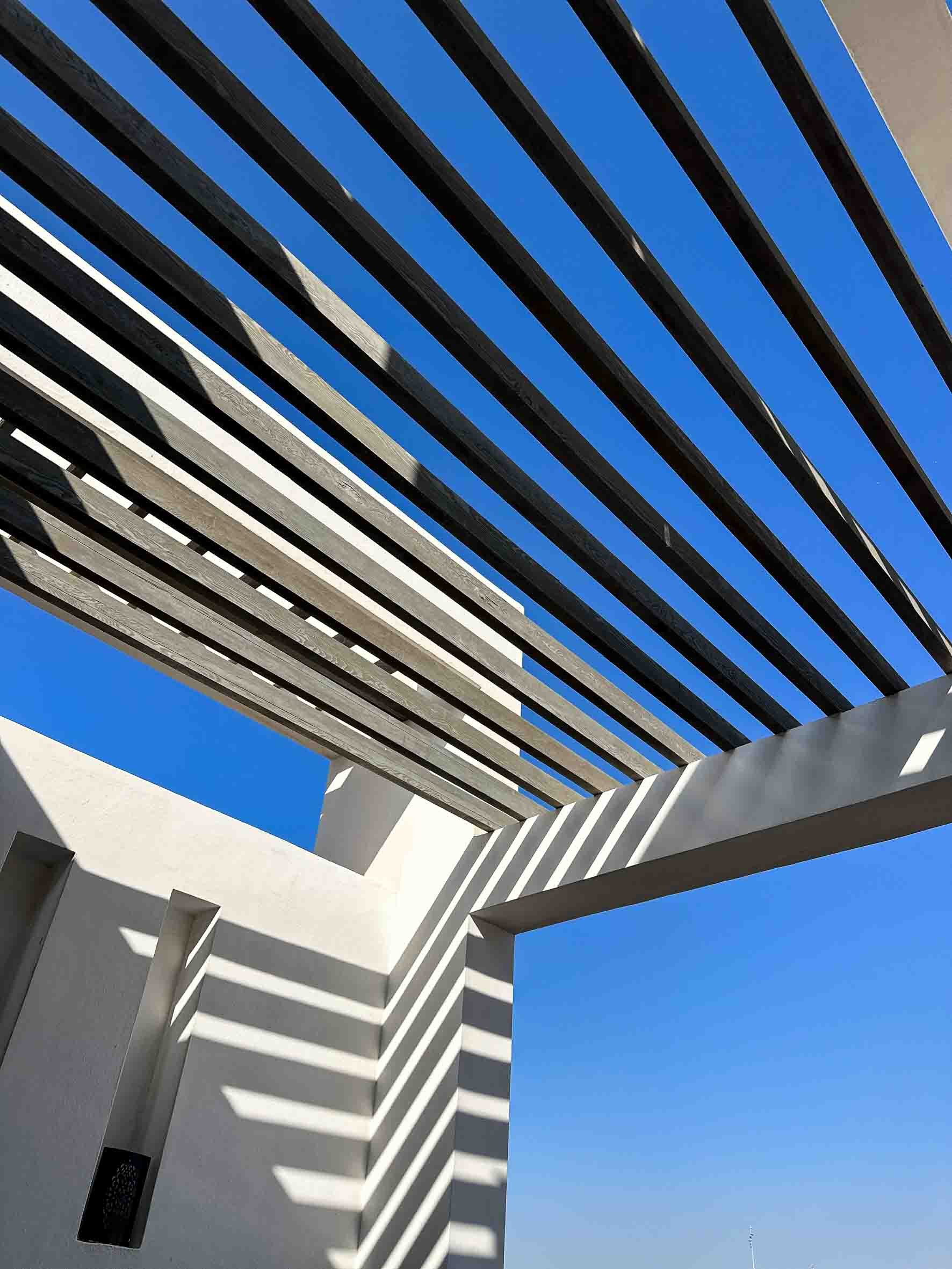 Architectural structure with white walls and horizontal wooden slats casting shadows against a clear blue sky.