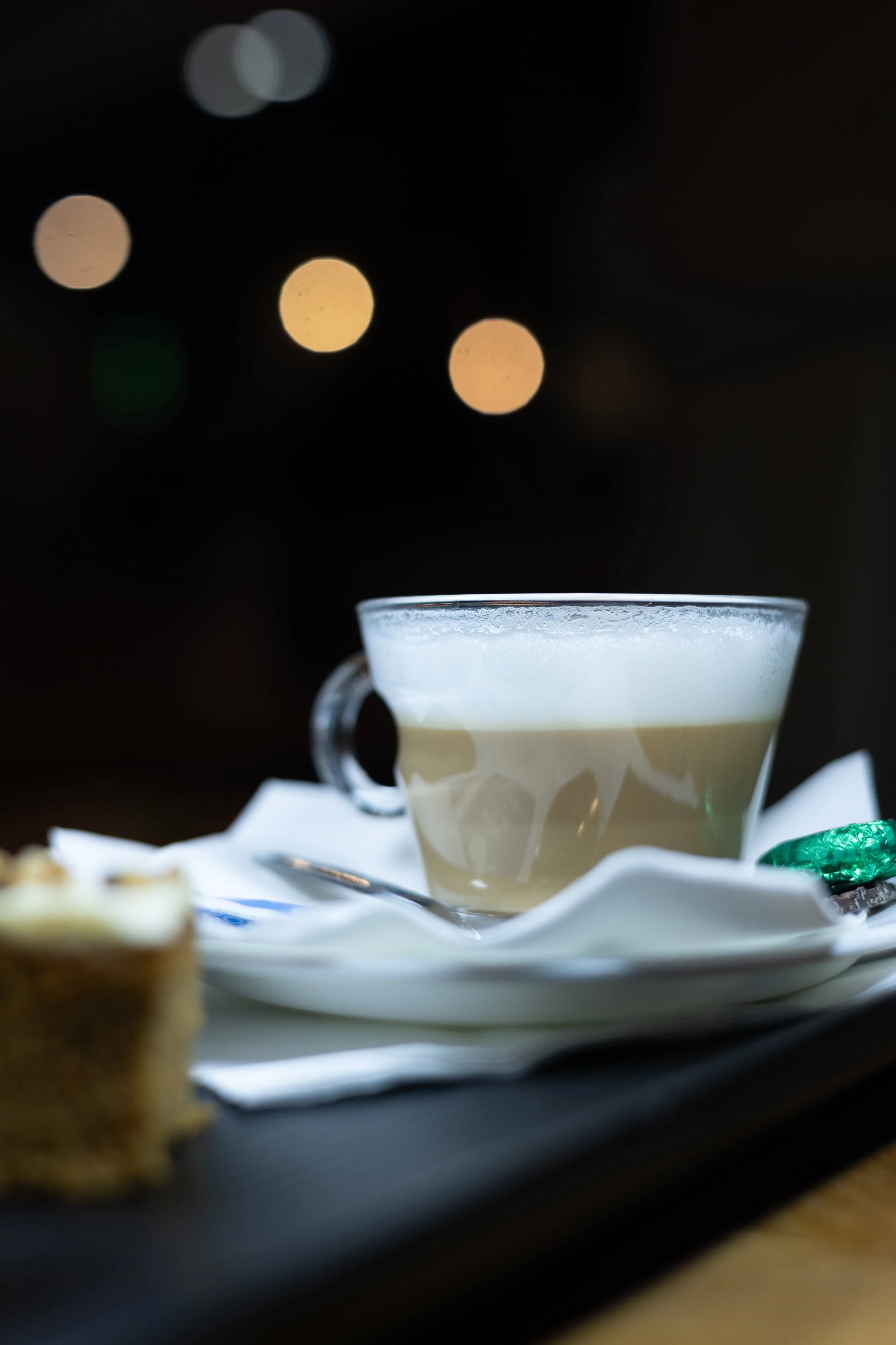 A cup of coffee with frothy top sitting on a saucer, with a slice of cake in the foreground, on a dark table with blurred lights in the background.