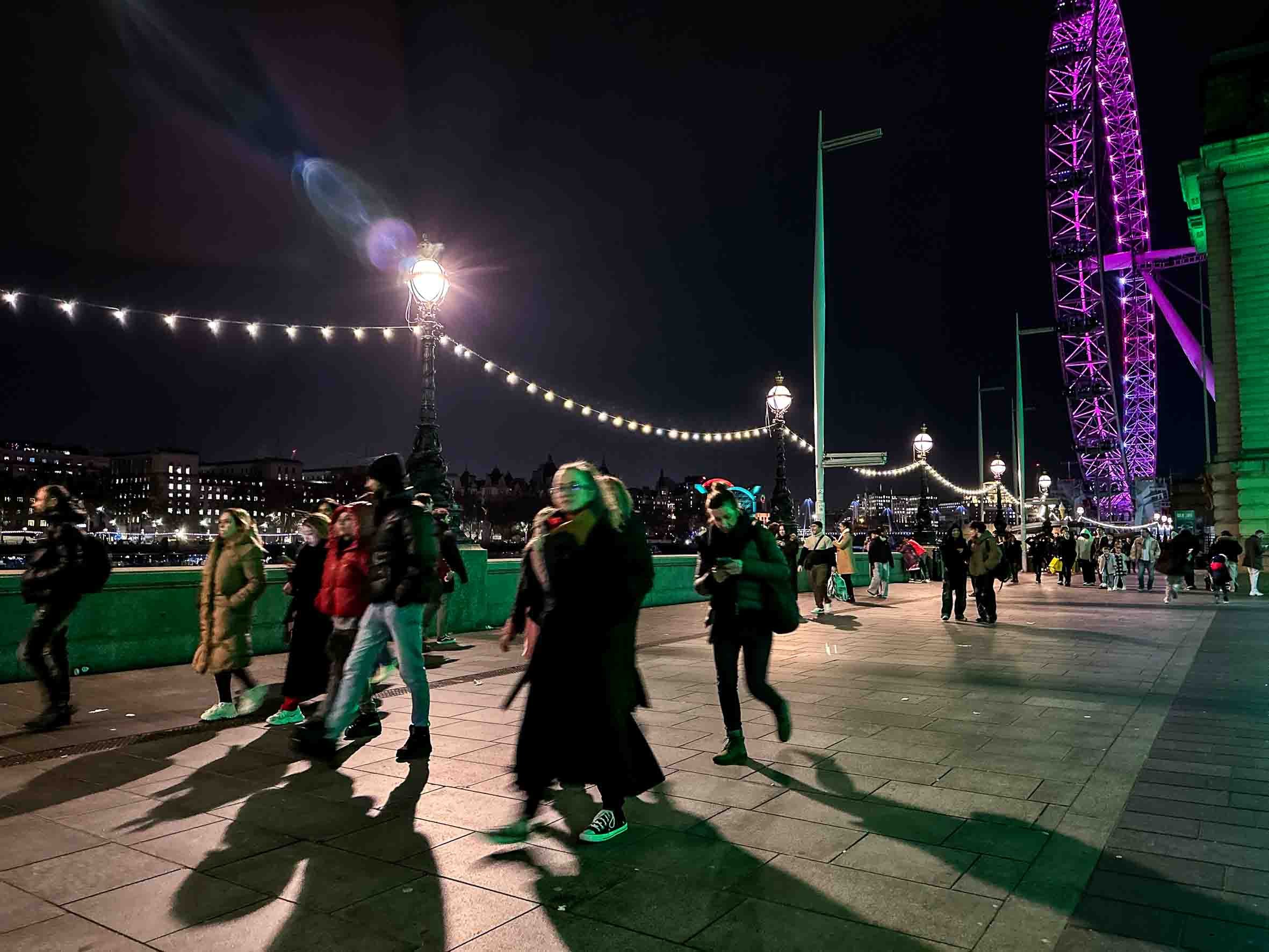 Night scene with people walking along a promenade next to a river, illuminated street lamps, and a large Ferris wheel lit with purple lights in the background.