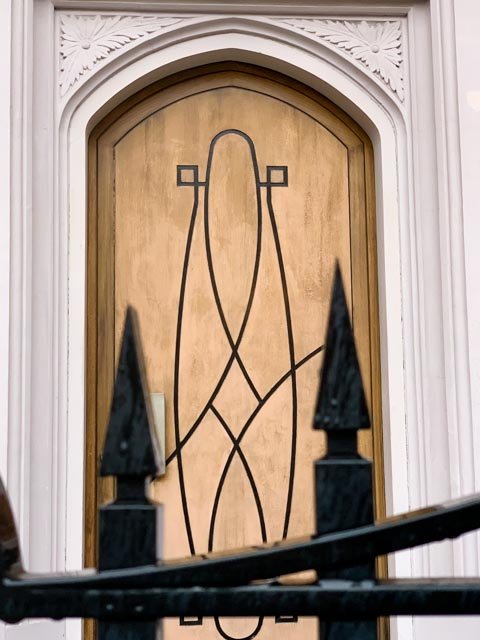 Wooden door with black metal decorative lines, framed by white molding with intricate carvings, partially obscured by black iron gate with pointed finials.