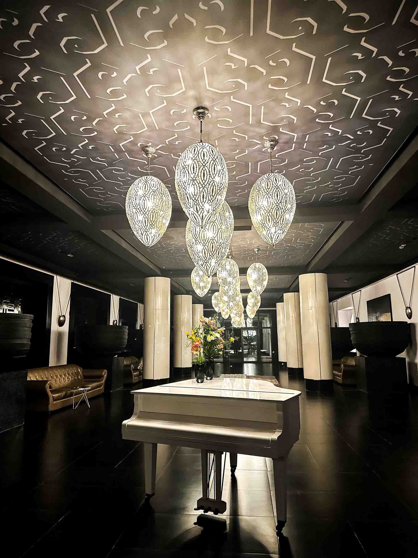 Elegant hotel lobby with a white grand piano, a flower arrangement on top, modern pendant lights hanging from a patterned ceiling, black and white pillars, and seating areas with leather couches.