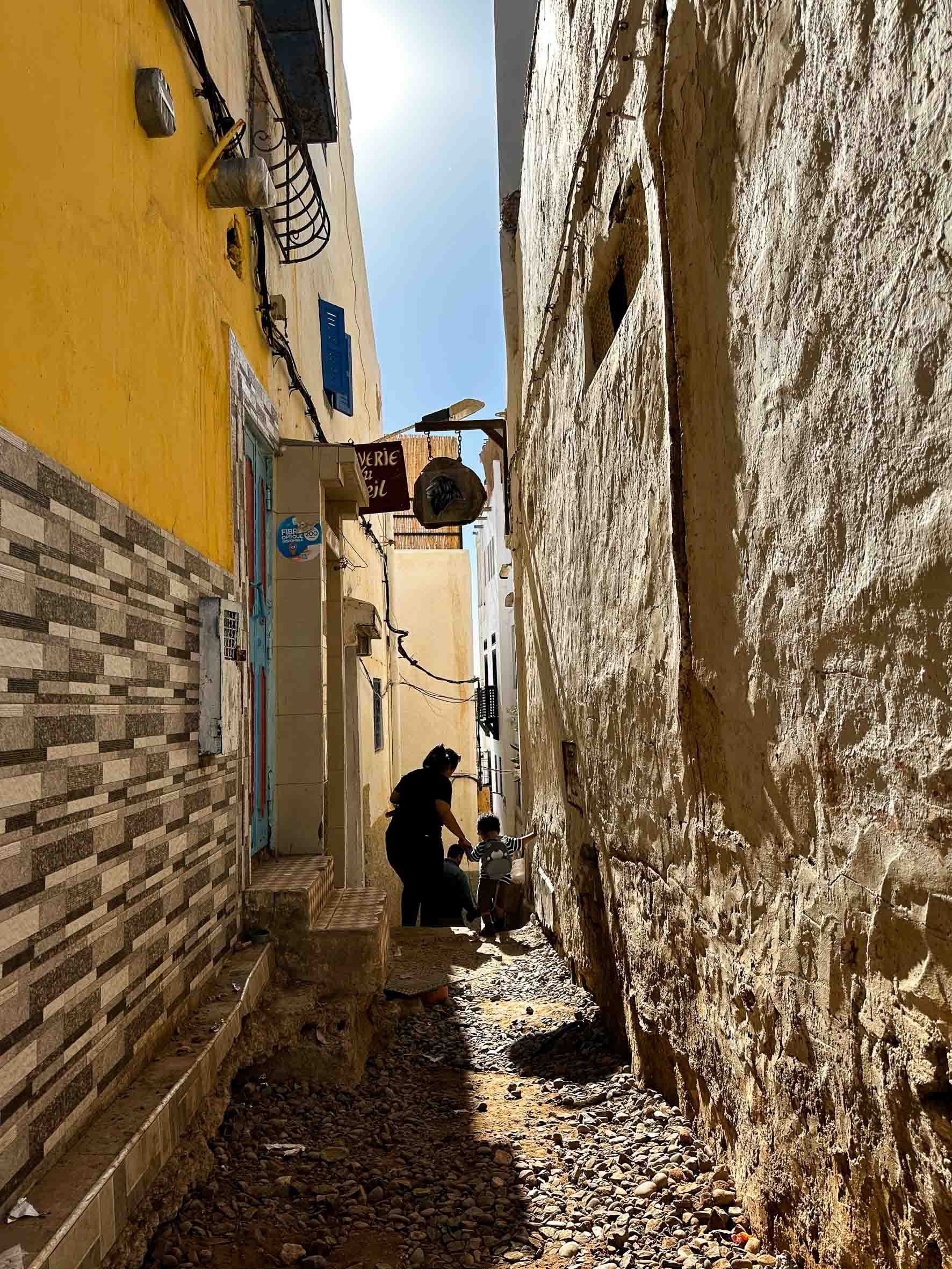 A narrow alleyway with a family walking through, with one adult and two children. The walls on either side are textured, with one wall painted yellow and the other with exposed stone. The ground is rocky and uneven, with some tiles and debris. There