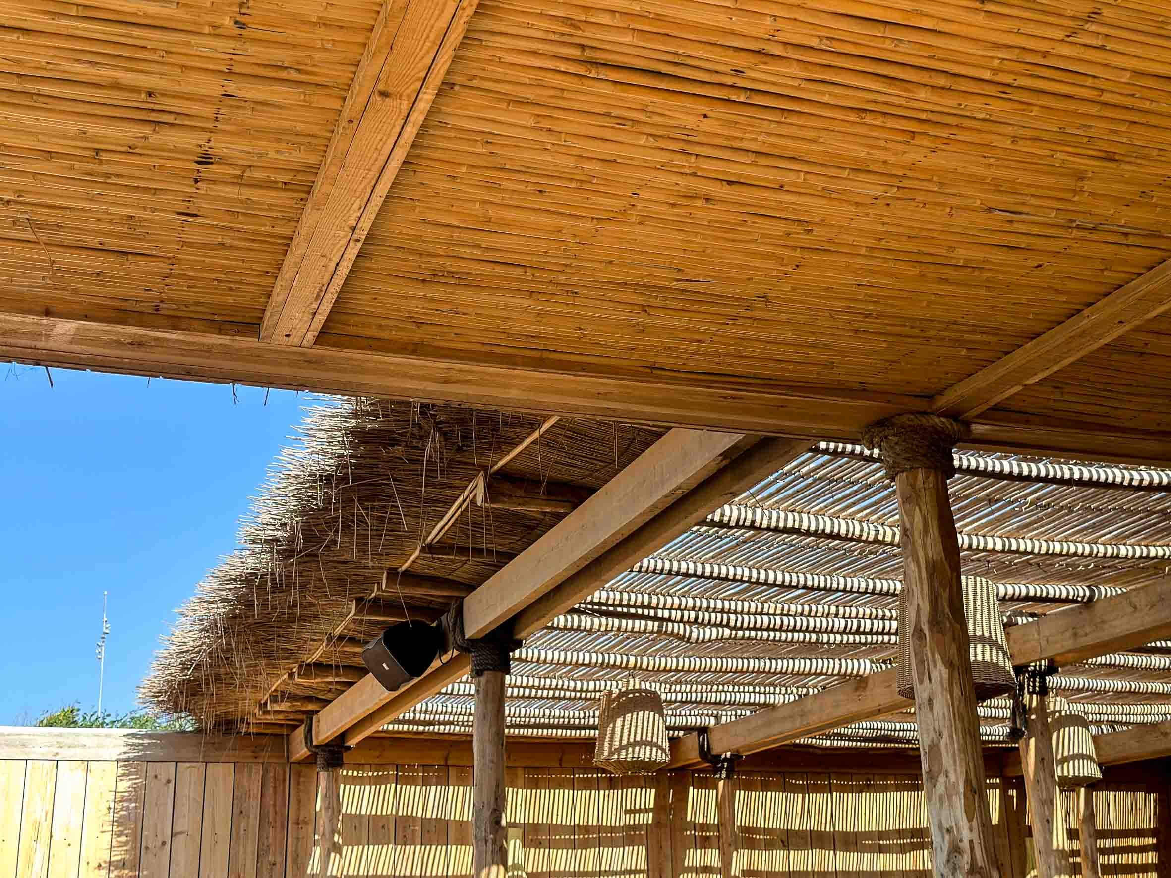 Part of an outdoor wooden structure with a bamboo roof, supported by wooden poles, under a bright blue sky.