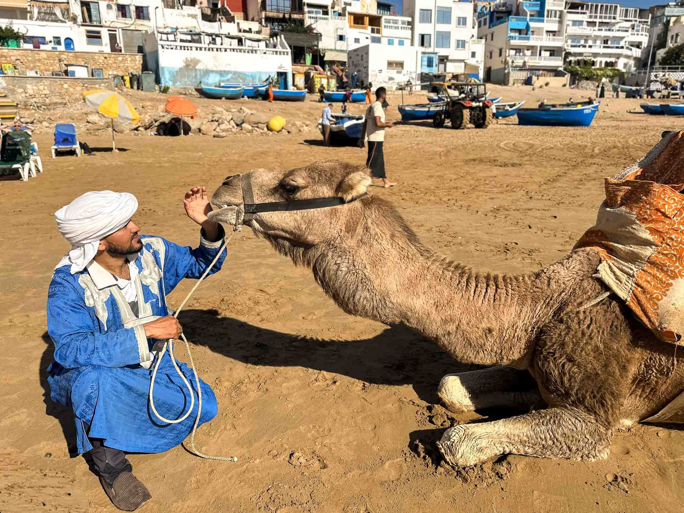 A man with a turban kneeling on a sandy beach, touching noses with a camel that has a pillow on its back, with buildings, boats, and people in the background.