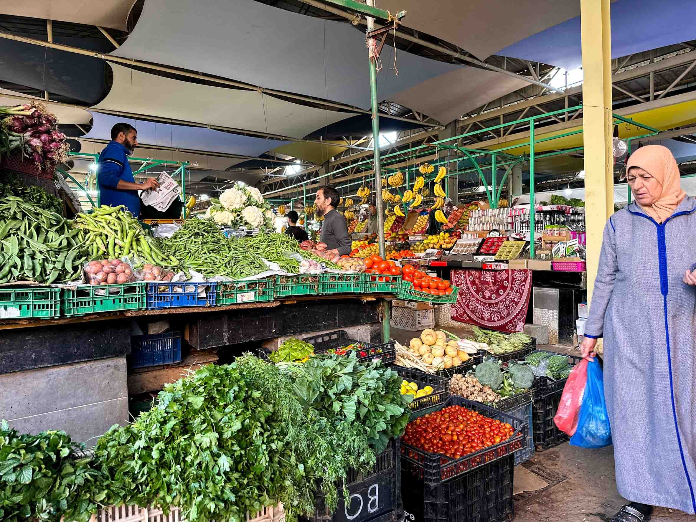 A bustling market stall with fresh vegetables, bananas, tomatoes, and other produce, with shoppers and vendors engaged in transactions under a colorful canopy.