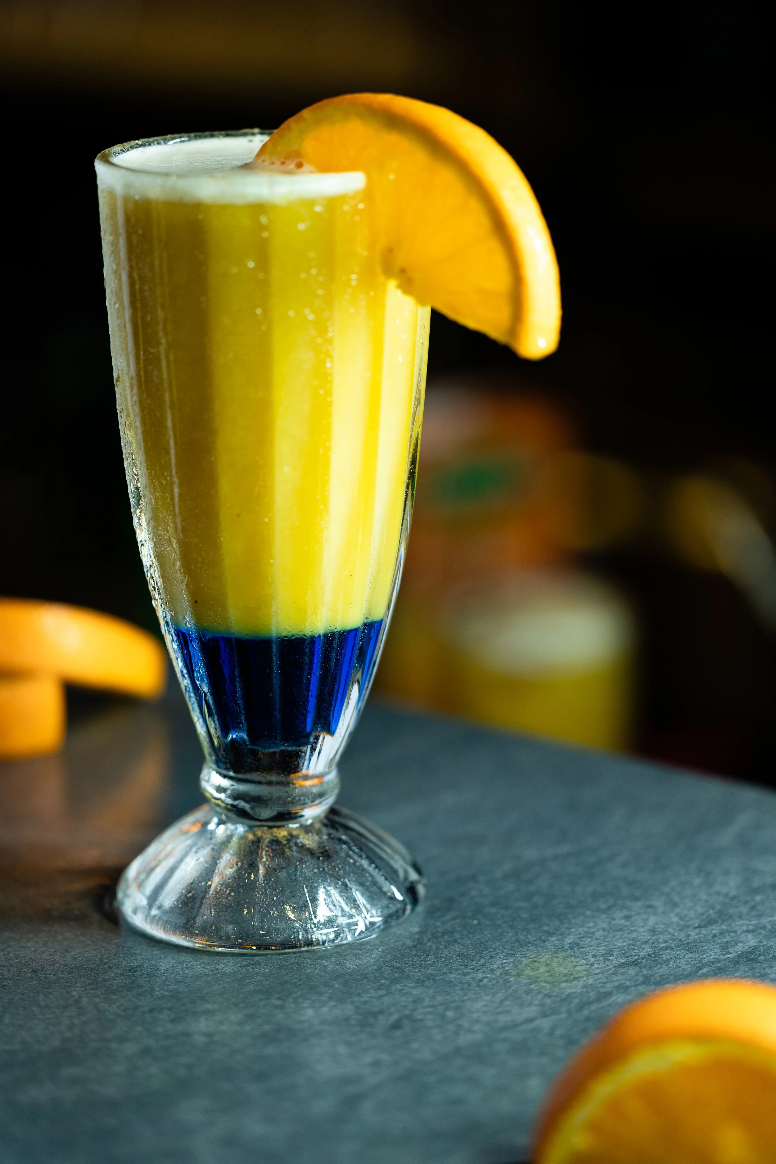 A tall glass with a yellow layered drink, garnished with an orange slice on the rim, on a dark surface.
