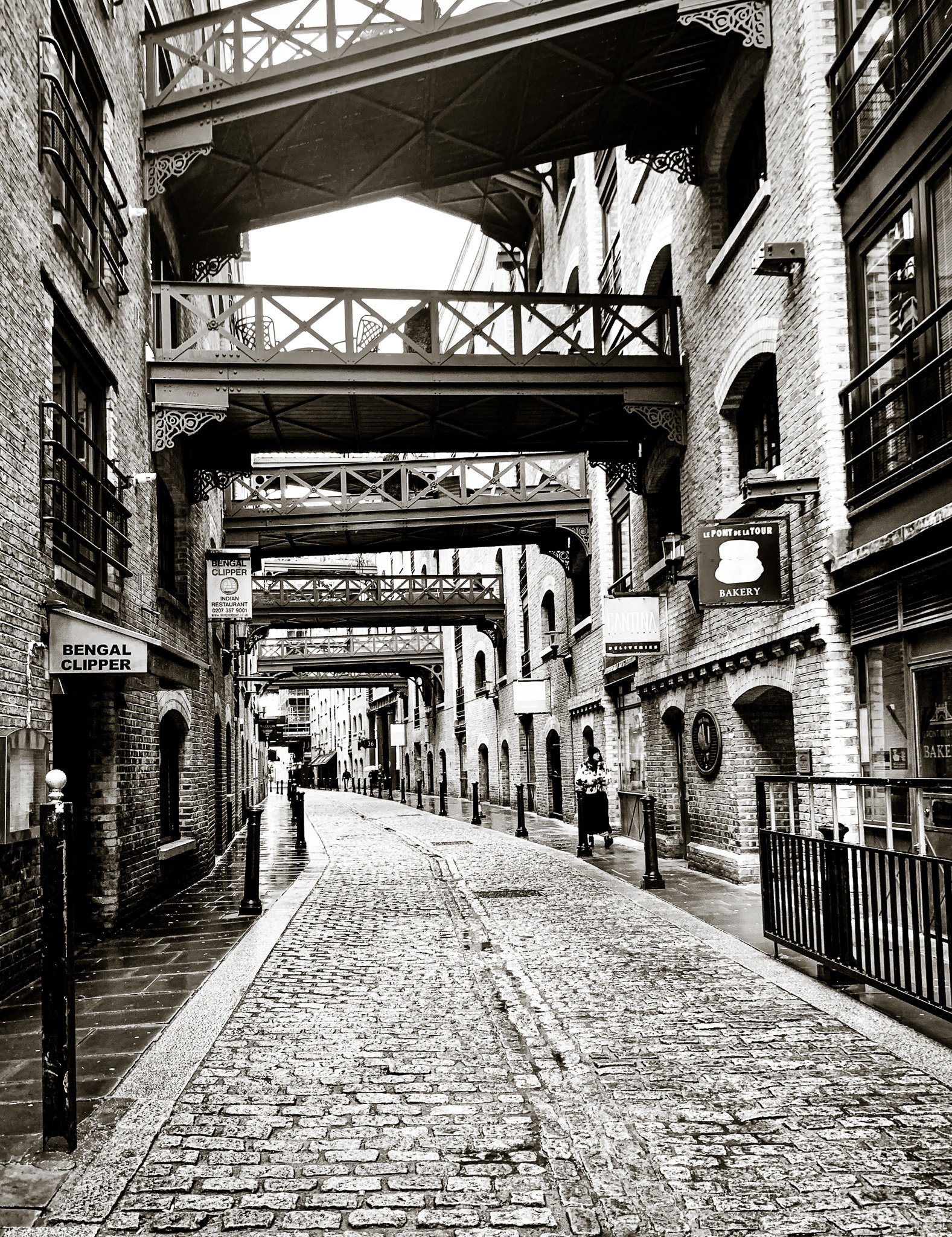 A black and white photo of a narrow cobblestone street with brick buildings on either side, featuring balconies and signs for shops and restaurants. overhead walkways connect the buildings.
