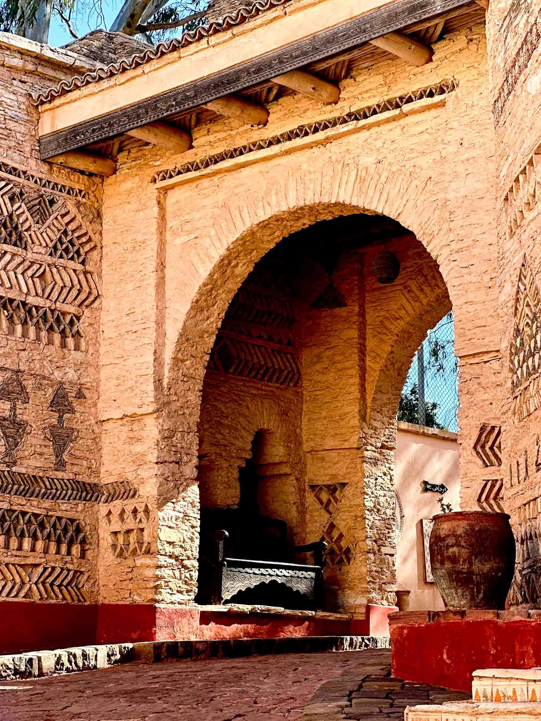 Ancient Moroccan-style entrance with intricate mud-brick walls, terracotta floor, and a large clay pot outside.