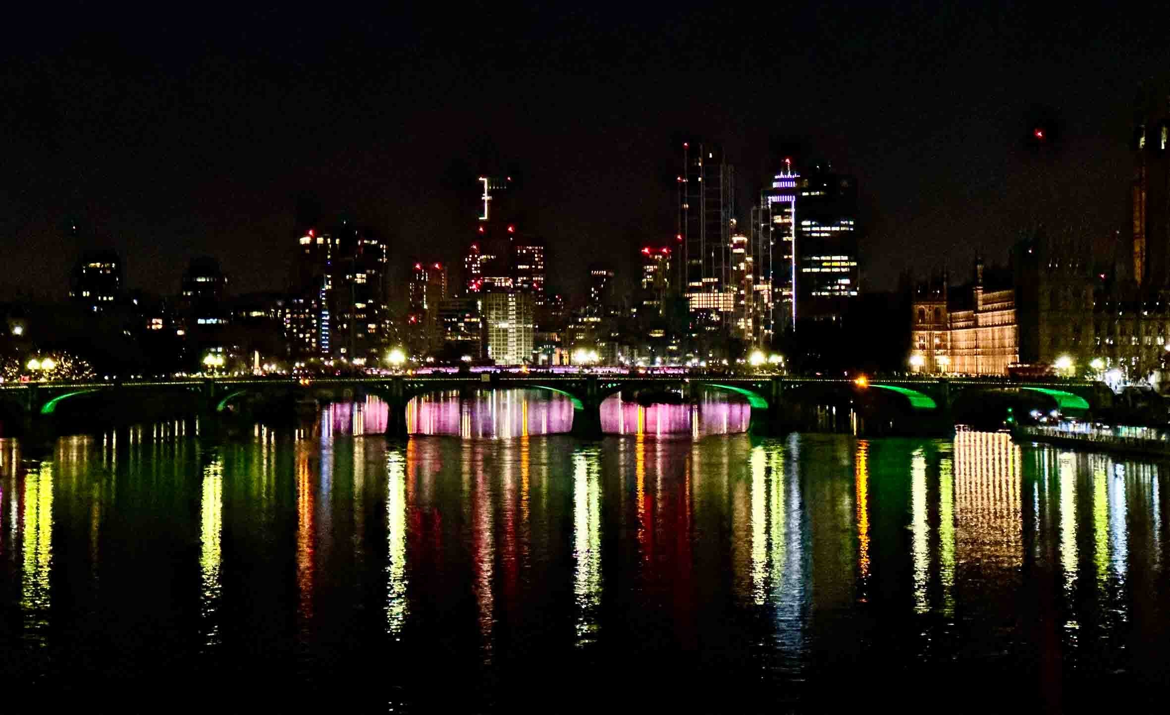Nighttime cityscape with illuminated buildings and a bridge reflected in the water.