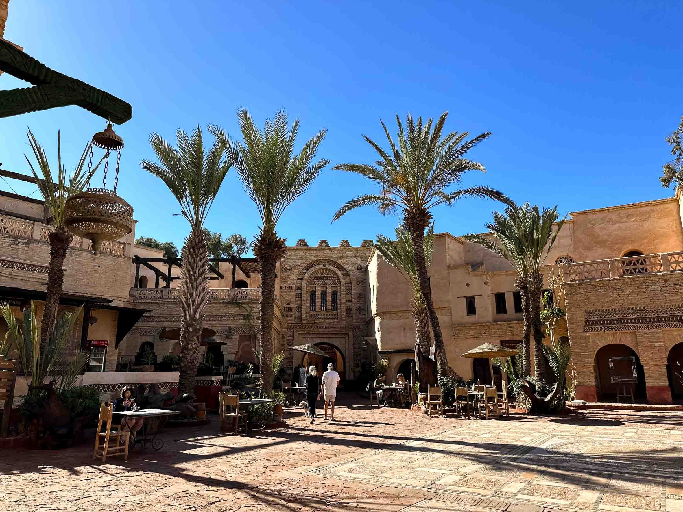 A sunny outdoor courtyard with palm trees, people sitting at tables, and historic-looking beige buildings with intricate facade details.