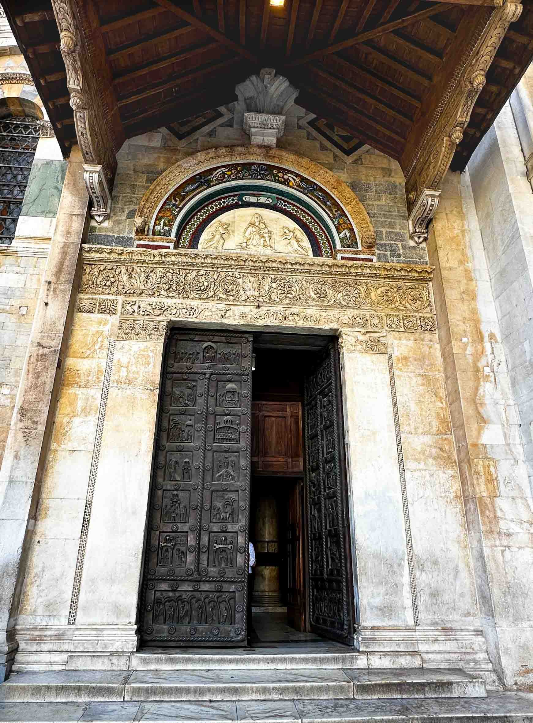 The entrance to a historic church or cathedral with large, detailed black doors, ornate stone carvings above the door, and religious artwork and mosaics set in an arched frame above the doorway.