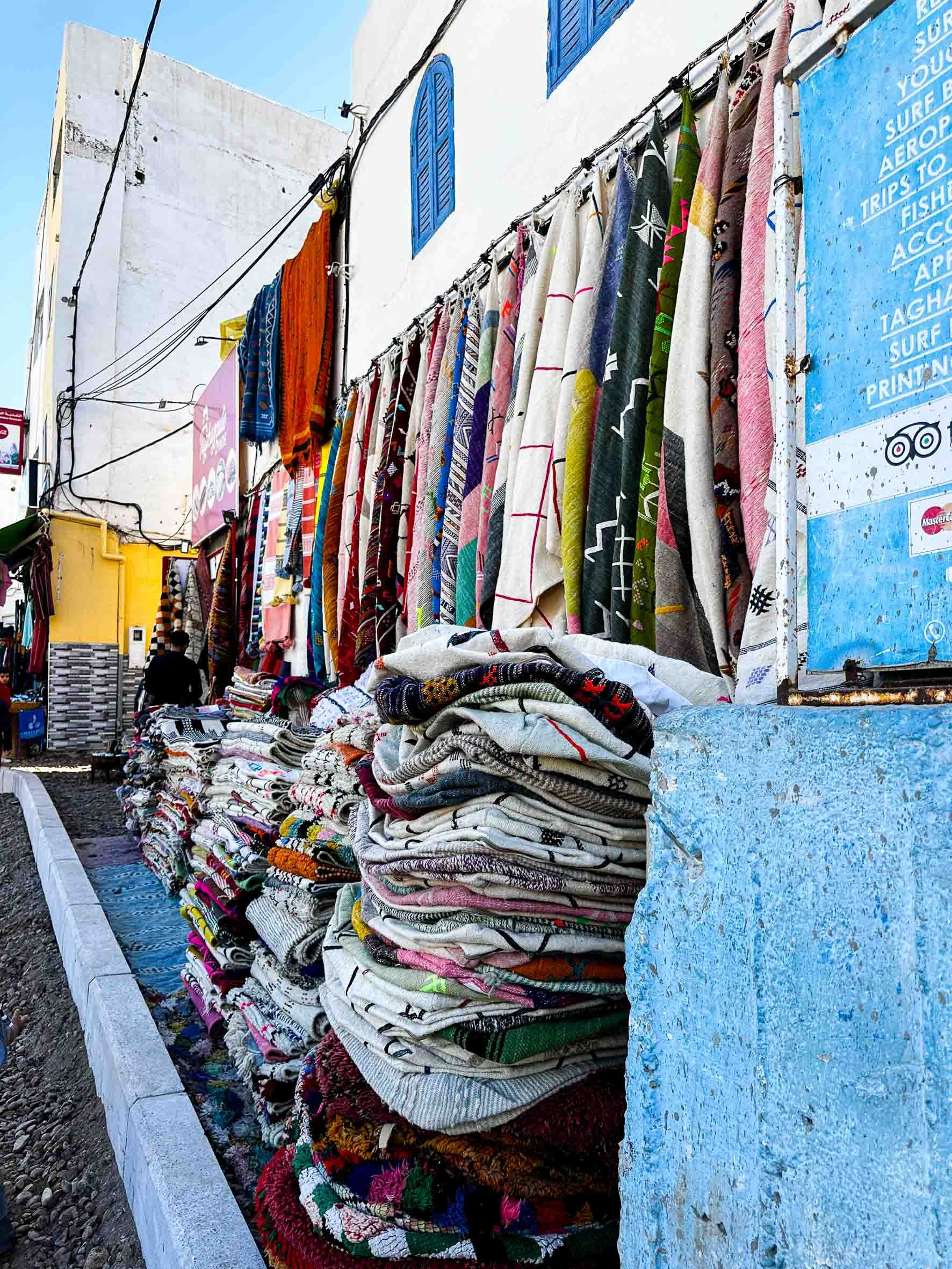 Stacks of colorful woven rugs and hanging textiles on display outside a shop with white and blue painted walls.