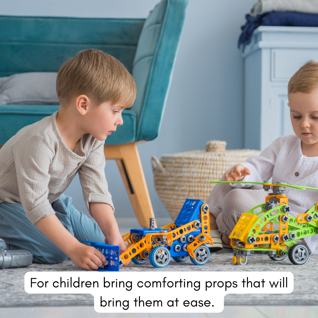 Two young children playing with colorful toy vehicles on a carpeted floor in a cozy room. One child is kneeling and the other sitting, focused on the toys. A teal sofa and white furniture are visible in the background.
