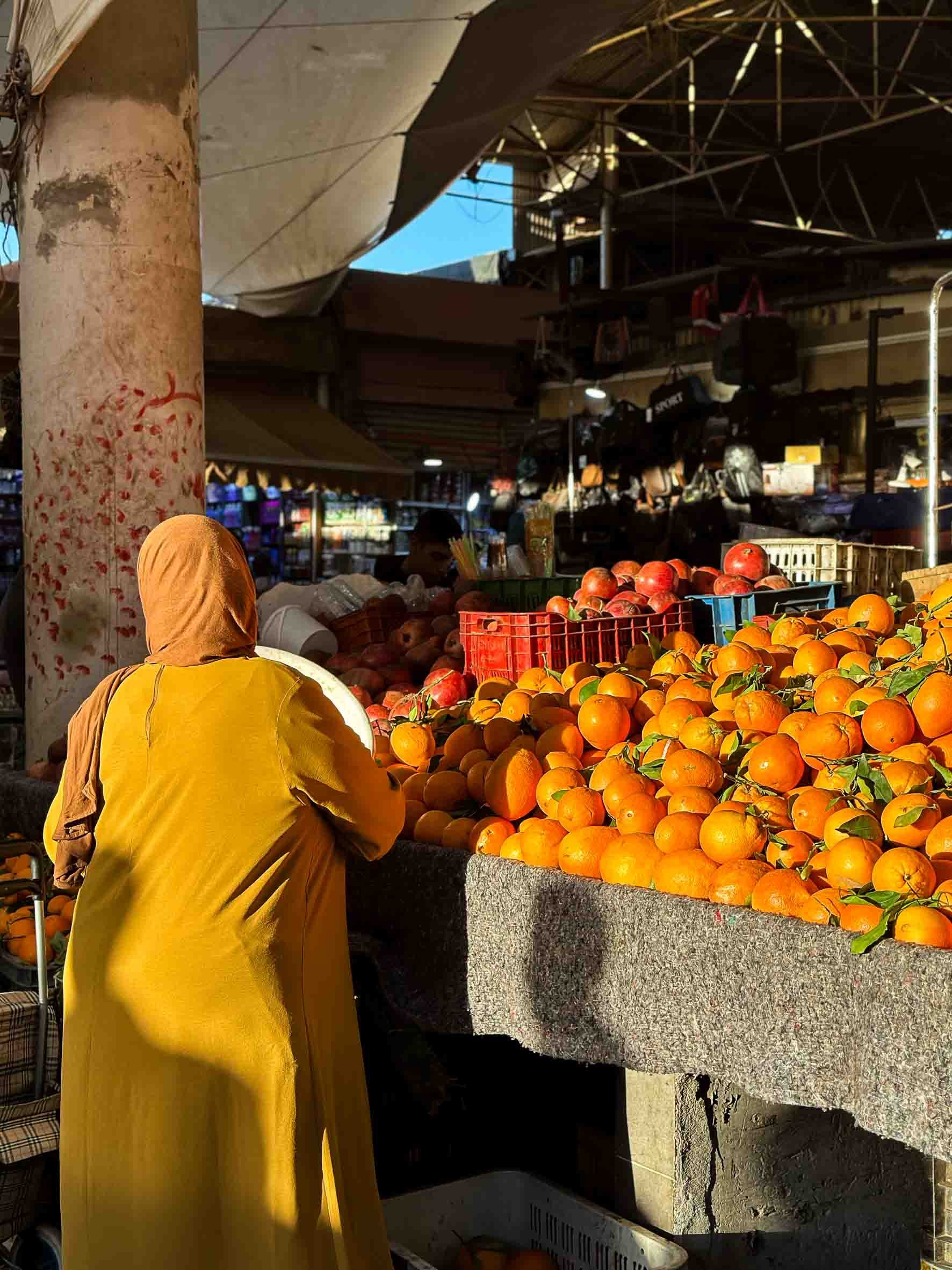 Person wearing a yellow cloak and peach-colored headscarf standing at a fruit stall with oranges, apples, and other fruits at an outdoor market during daytime.