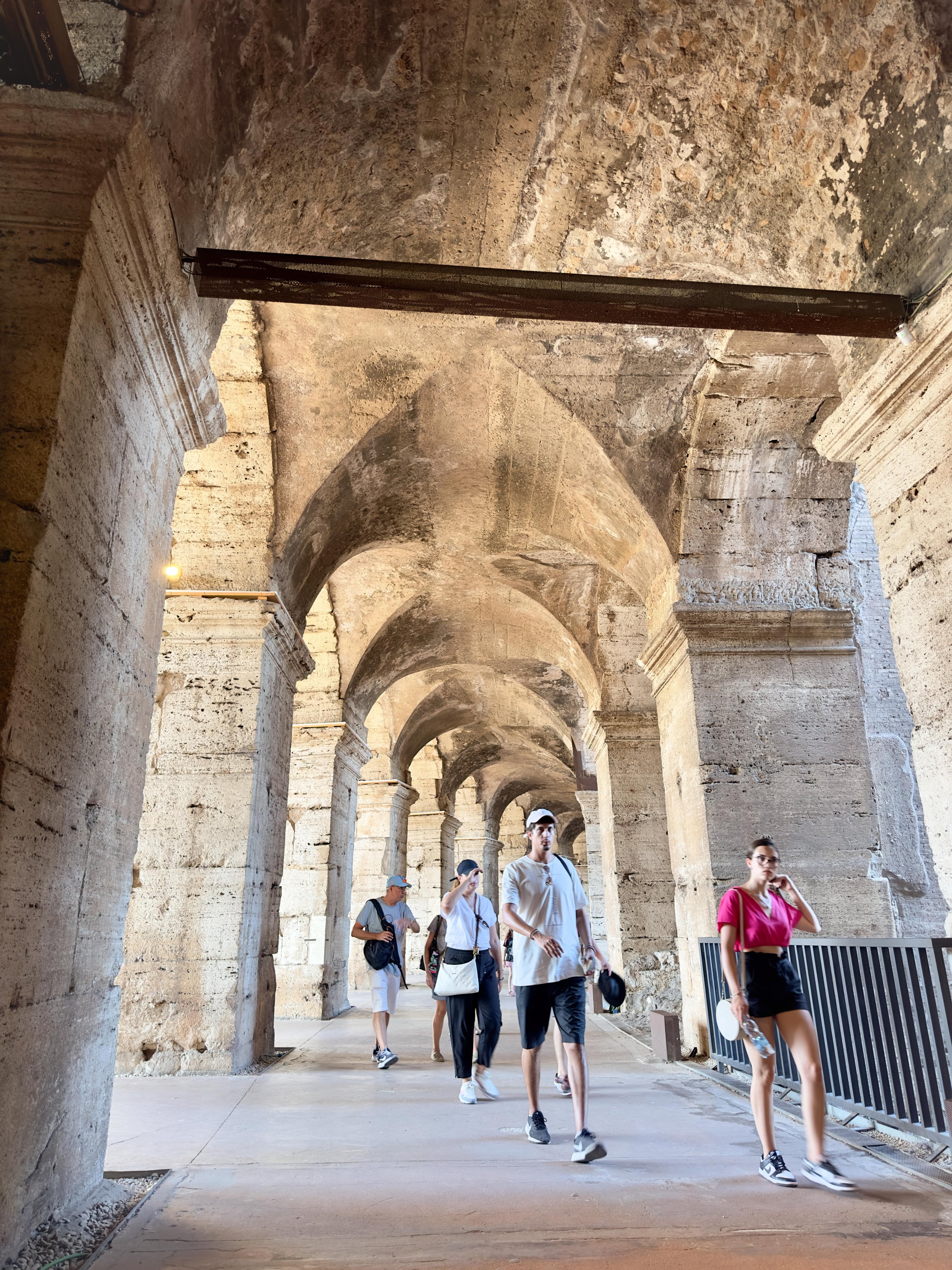 Tourists walking through an ancient stone corridor with arches, some taking photos, in a historical site or ruin.