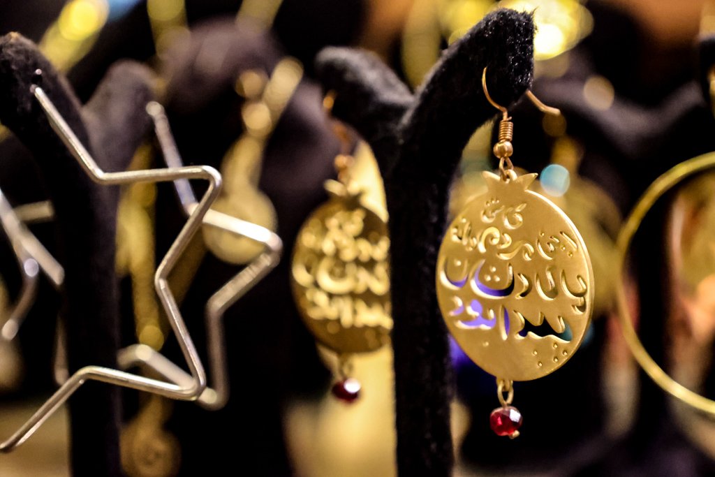 Gold earrings with Arabic calligraphy hanging on a black display stand, surrounded by other jewelry pieces.