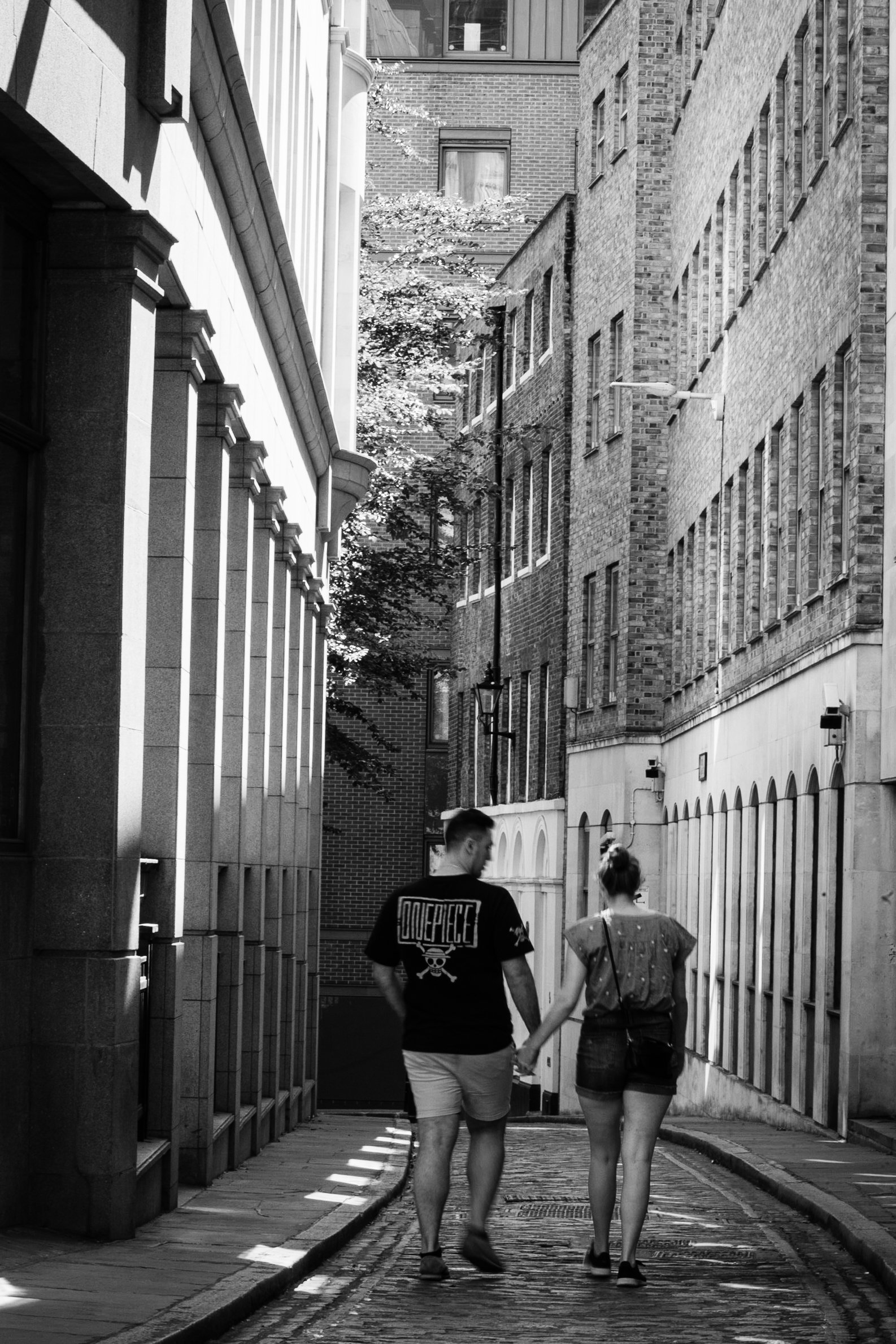 A black and white photo of a couple walking hand-in-hand down a narrow, cobblestone alleyway surrounded by tall brick and stone buildings.