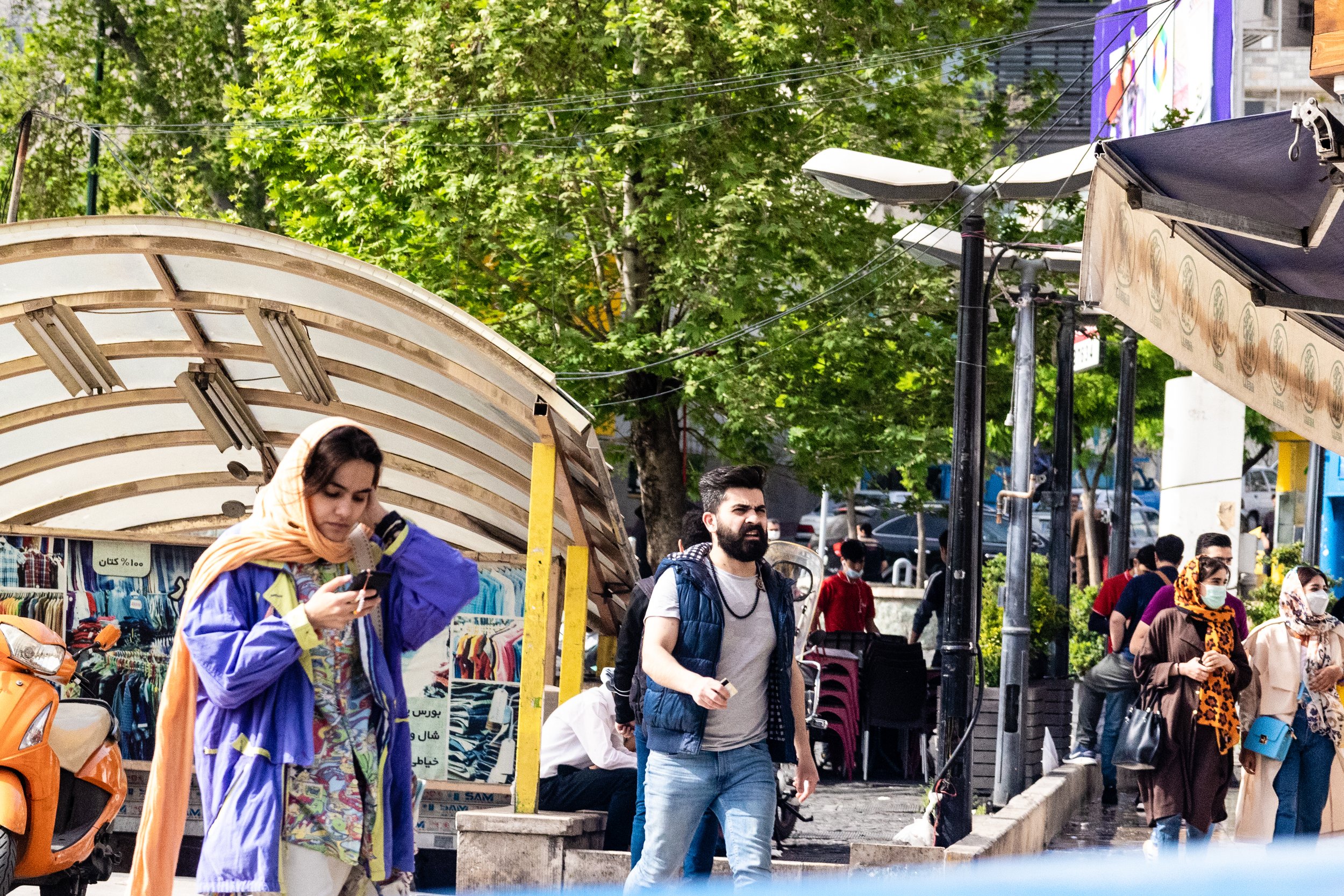 People walking on a city sidewalk with trees in the background and market stalls along the street. Some individuals are wearing masks; one woman is looking at her phone, and a man with a backpack is walking. Streetlights and power lines are visible.