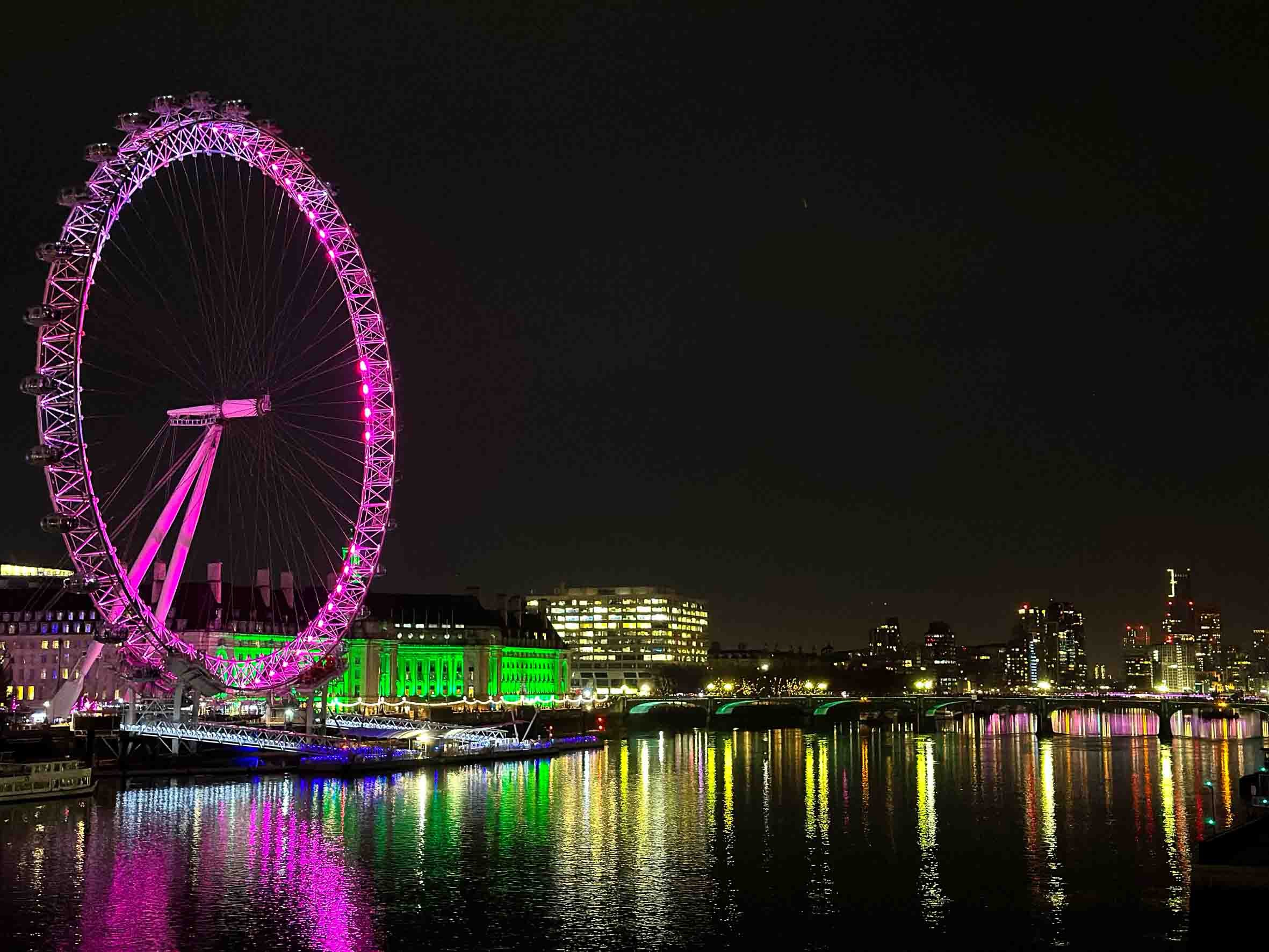 Night view of a brightly lit cityscape featuring a large illuminated pink Ferris wheel near a river, with colorful reflections on the water. Buildings with lights are visible in the background.