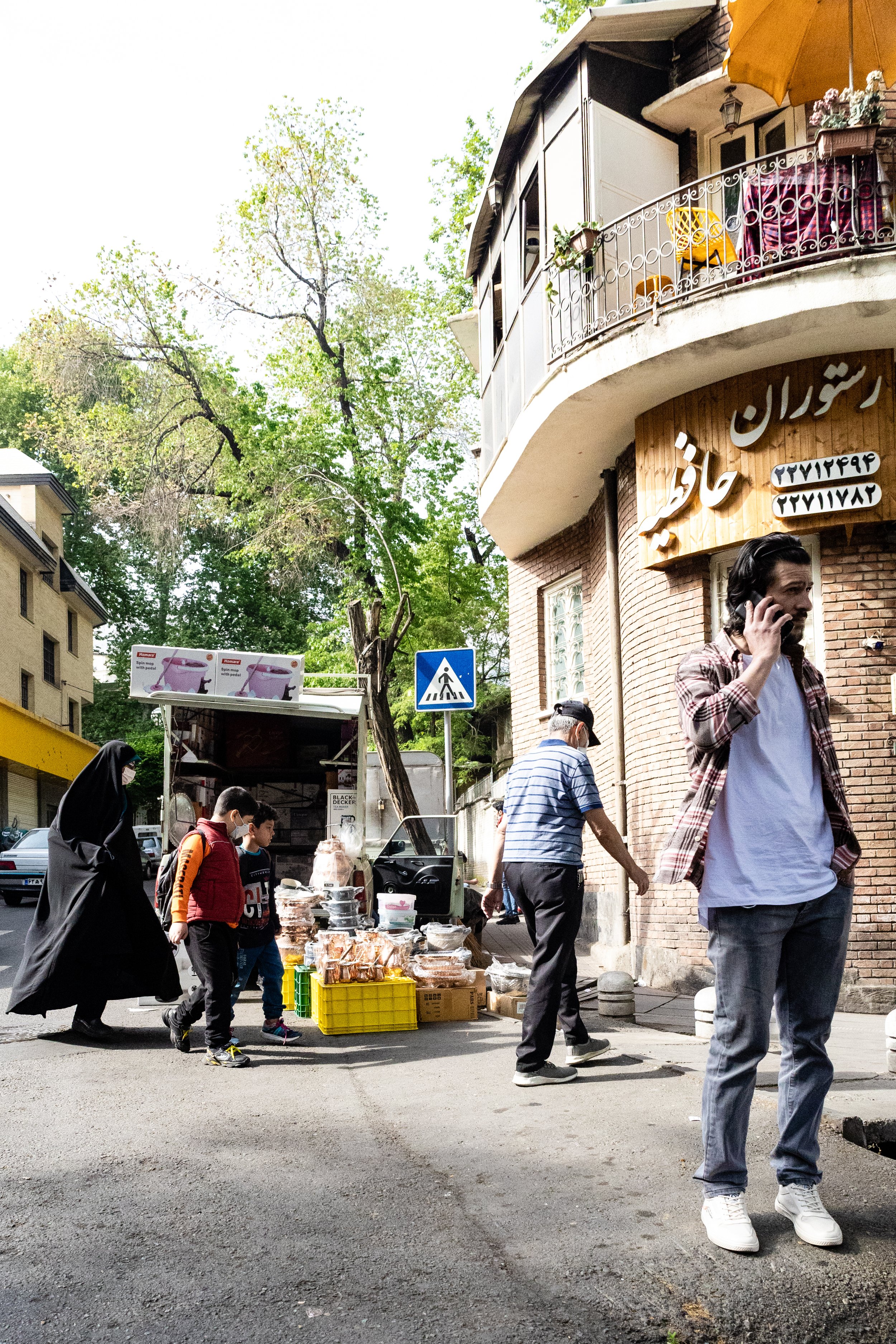 People walking along a street with a food stand and a man talking on his phone. There are trees, buildings, and a pedestrian crosswalk sign in the scene.