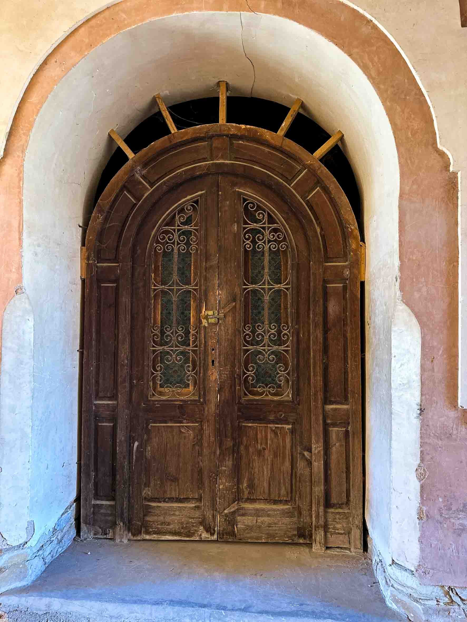 Old wooden double door with decorative ironwork and glass panels, framed by a rounded archway with wooden beams above.