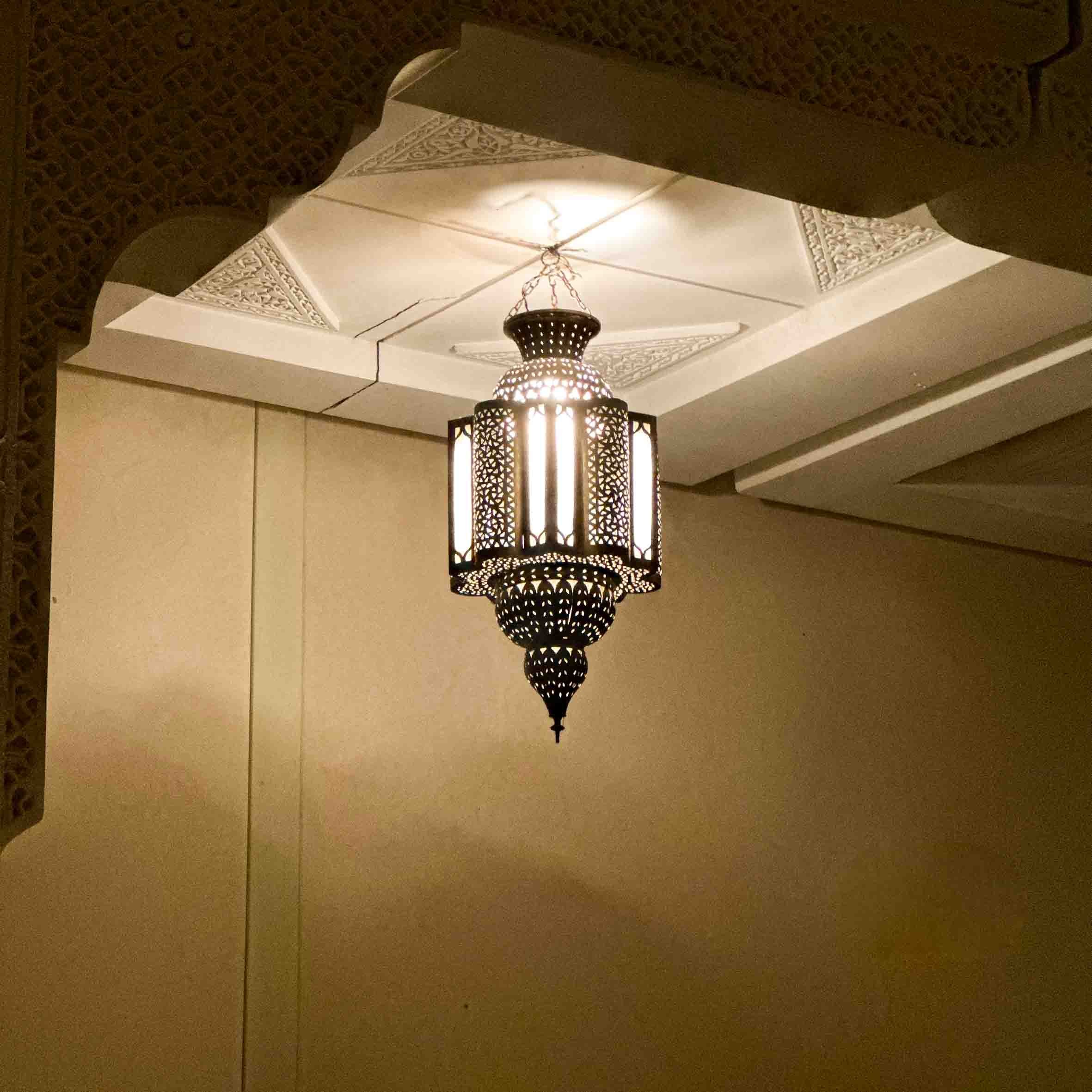 A decorative hanging lantern with intricate cutouts, emitting light, suspended from a ceiling with ornate molding.