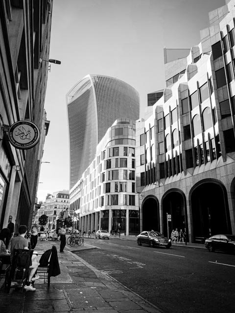 Black and white photo of a city street with modern buildings, cars, and people walking, with the Walkie Talkie building in the background.