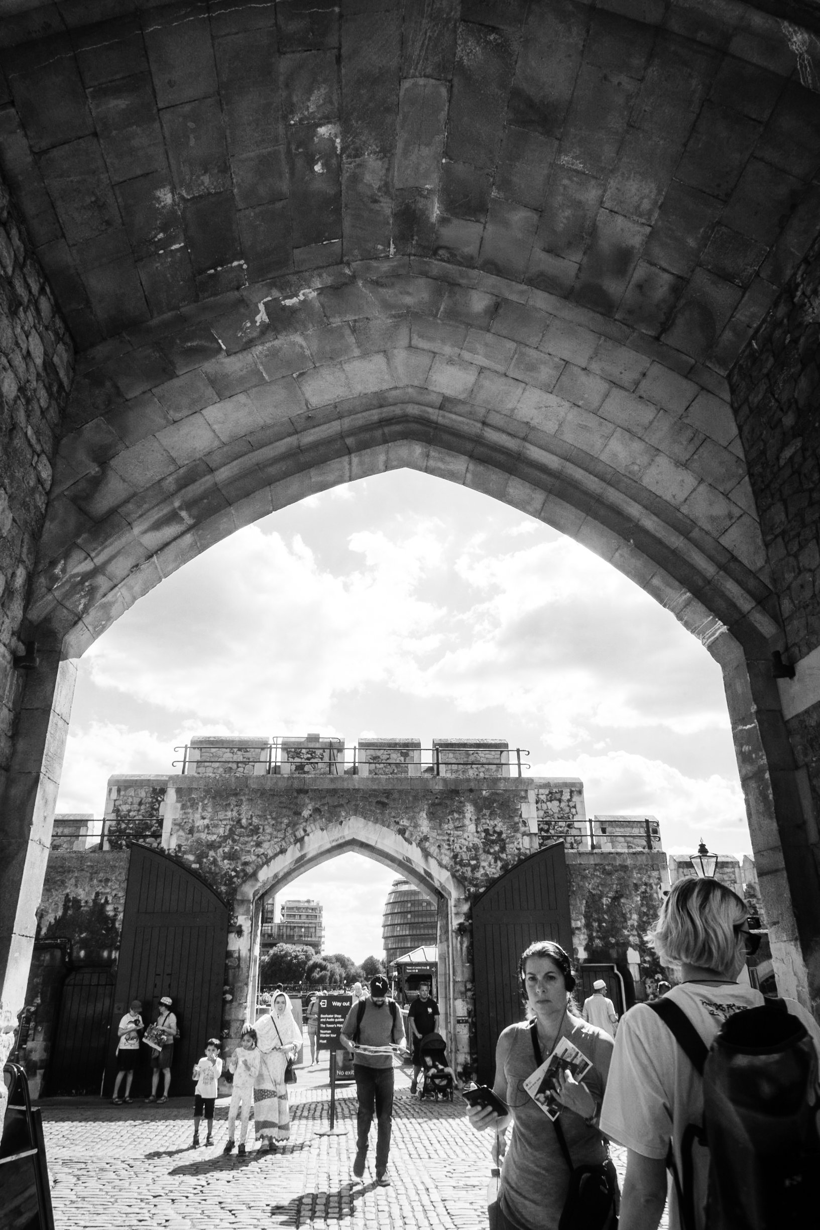 People walking out of an old stone city gate with a cobblestone street in front, some carrying backpacks and pamphlets, under a stone arch with a view of modern buildings and a cloudy sky in the background.