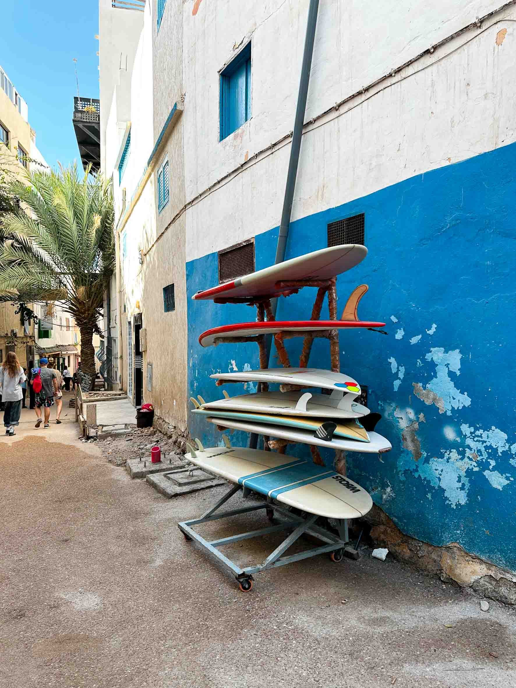 A rack of surfboards leaning against a weathered blue and white wall in a narrow street, with pedestrians and a palm tree visible in the background.