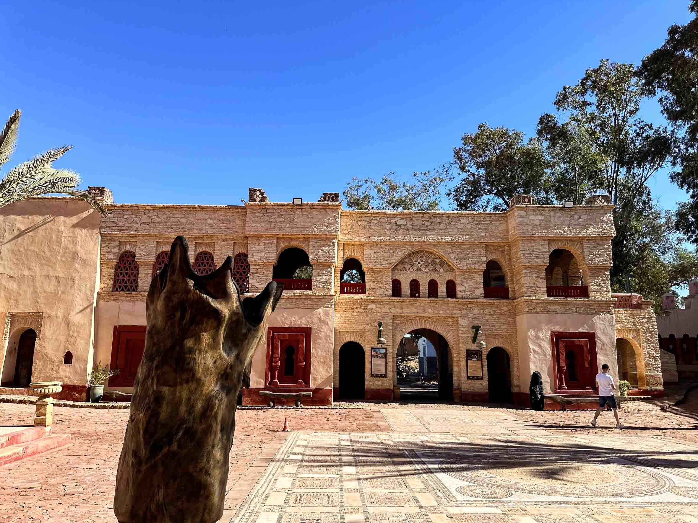 A person stands in front of a historic beige stone building with multiple arches and small windows, surrounded by trees, under a clear blue sky.