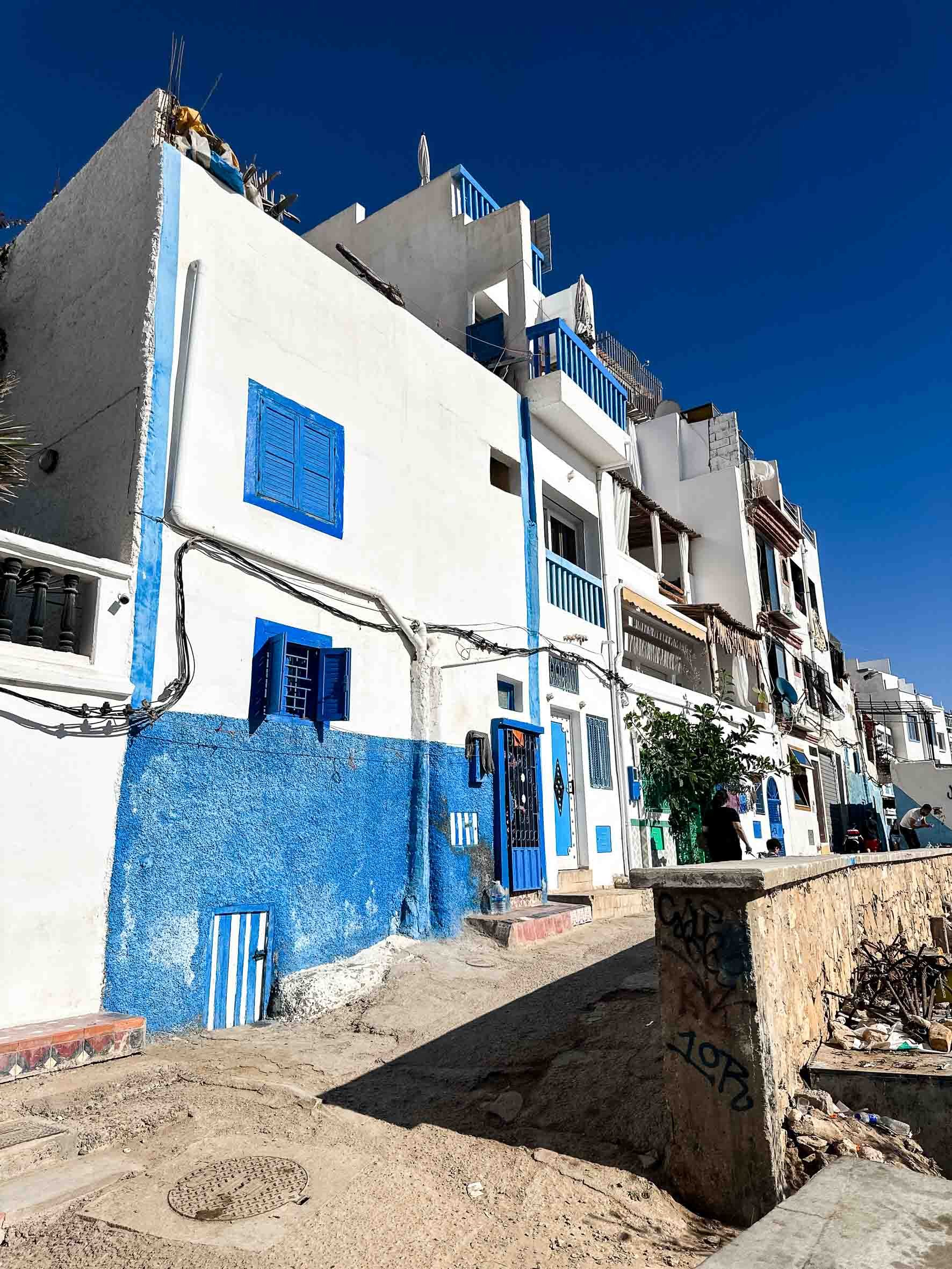 White buildings with blue accents and doors along a dirt street under a clear blue sky.