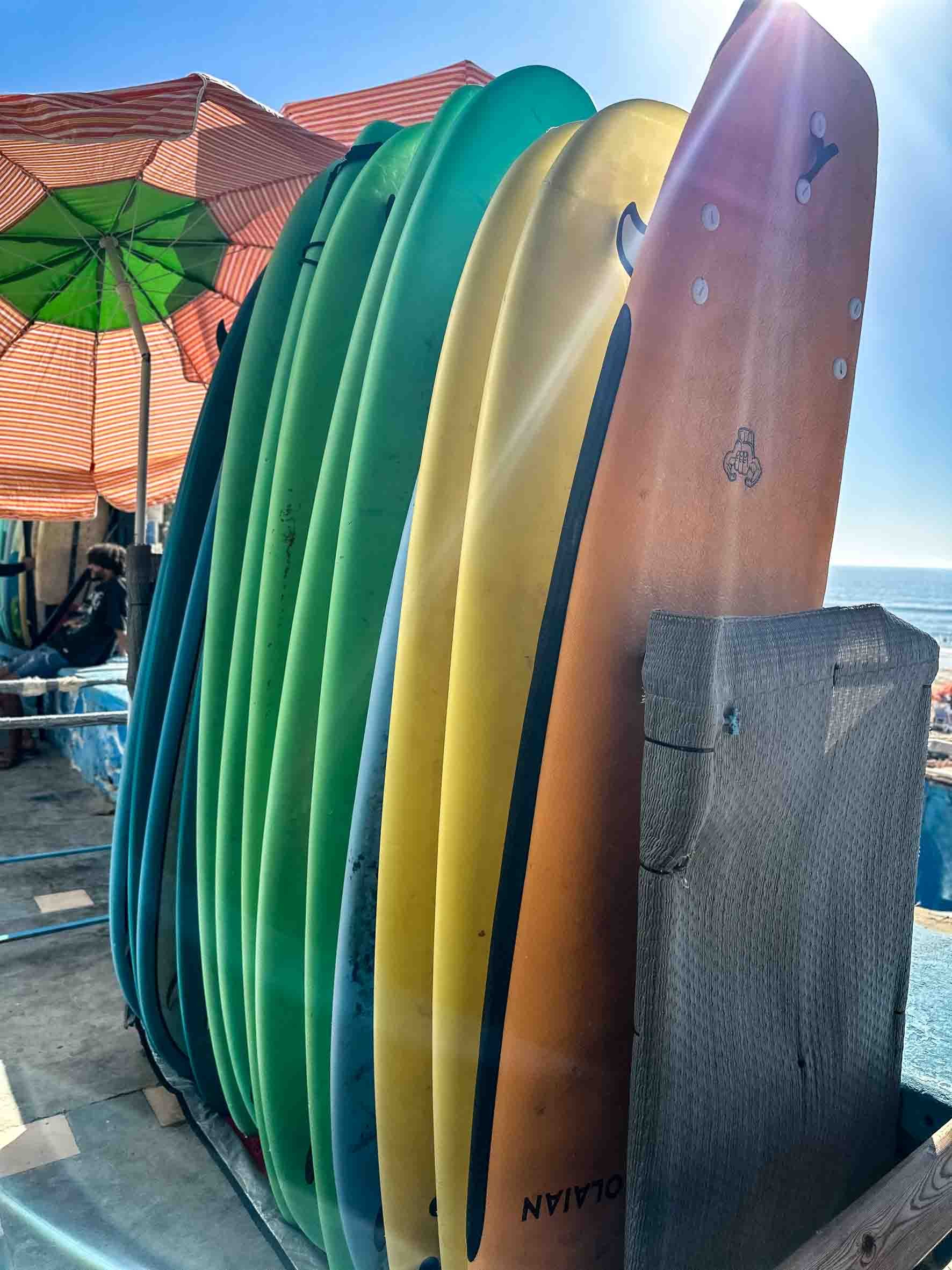 Stacked colorful surfboards standing upright on a beach, with umbrellas and the ocean in the background.