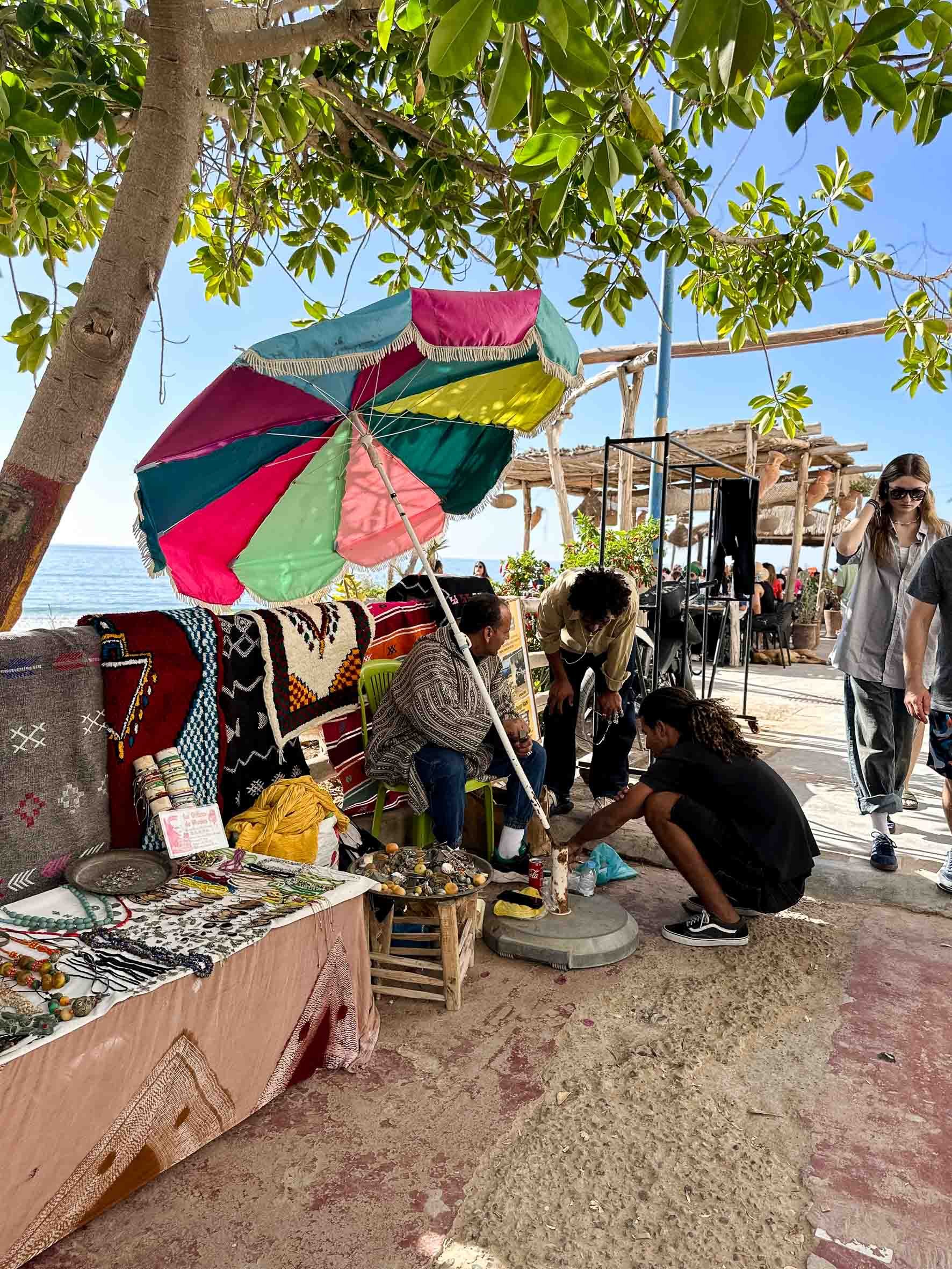 Vendors at an outdoor market selling jewelry and crafts beneath a colorful umbrella, near the beach with people walking and sitting in the background.