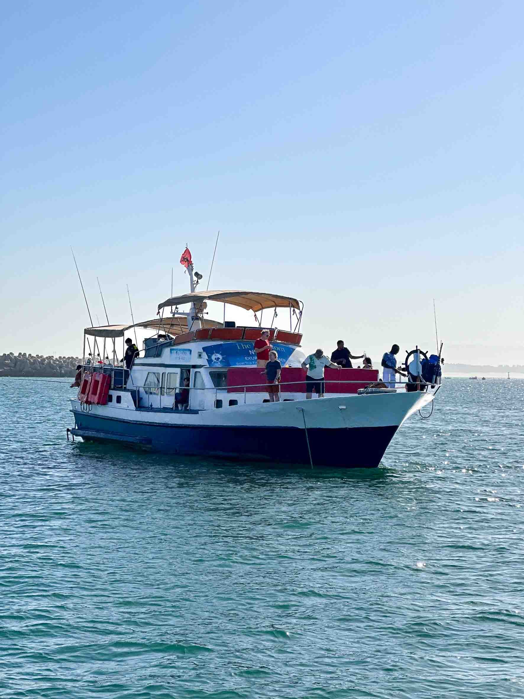 A group of people on a boat with a white and dark blue hull, sailing on the water during daytime, with a breakwater or rocky shoreline in the background.