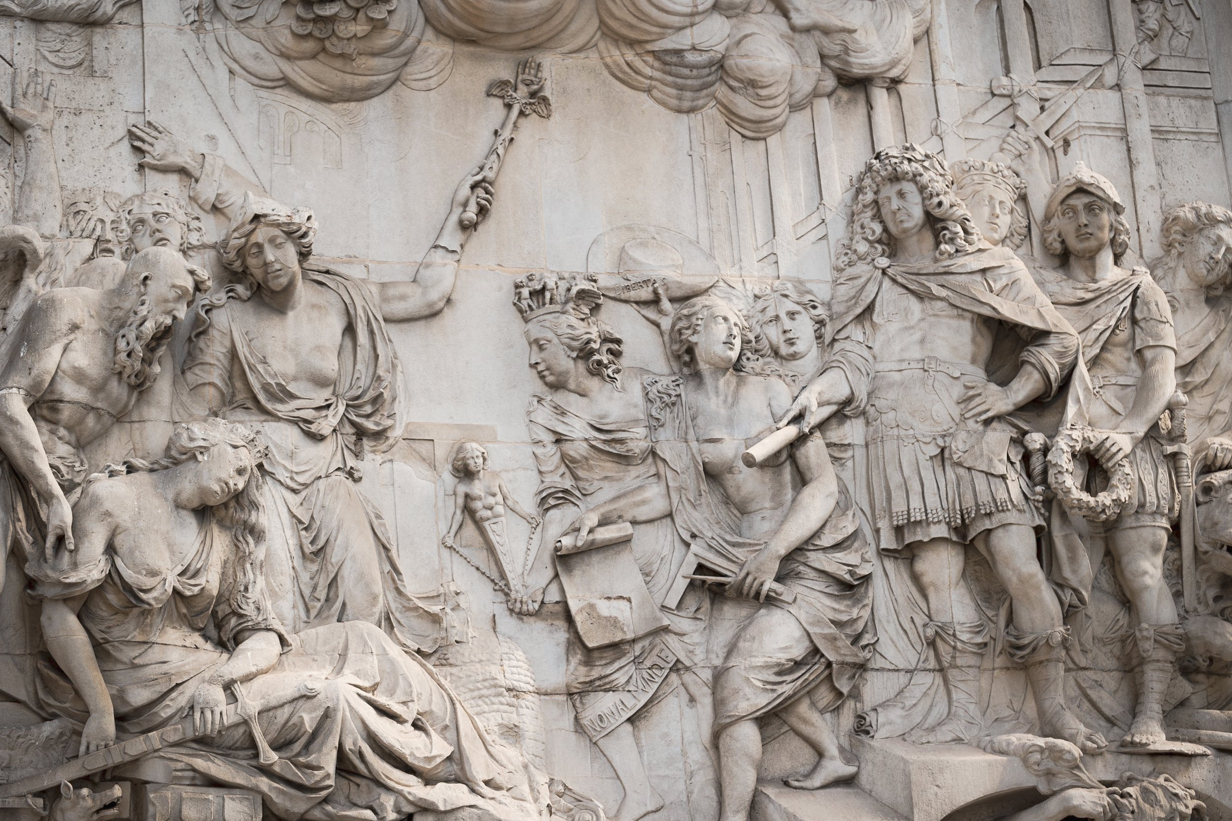 Detailed stone bas-relief sculpture depicting soldiers, a woman holding a scroll, and other figures, likely illustrating a historical or mythological scene.