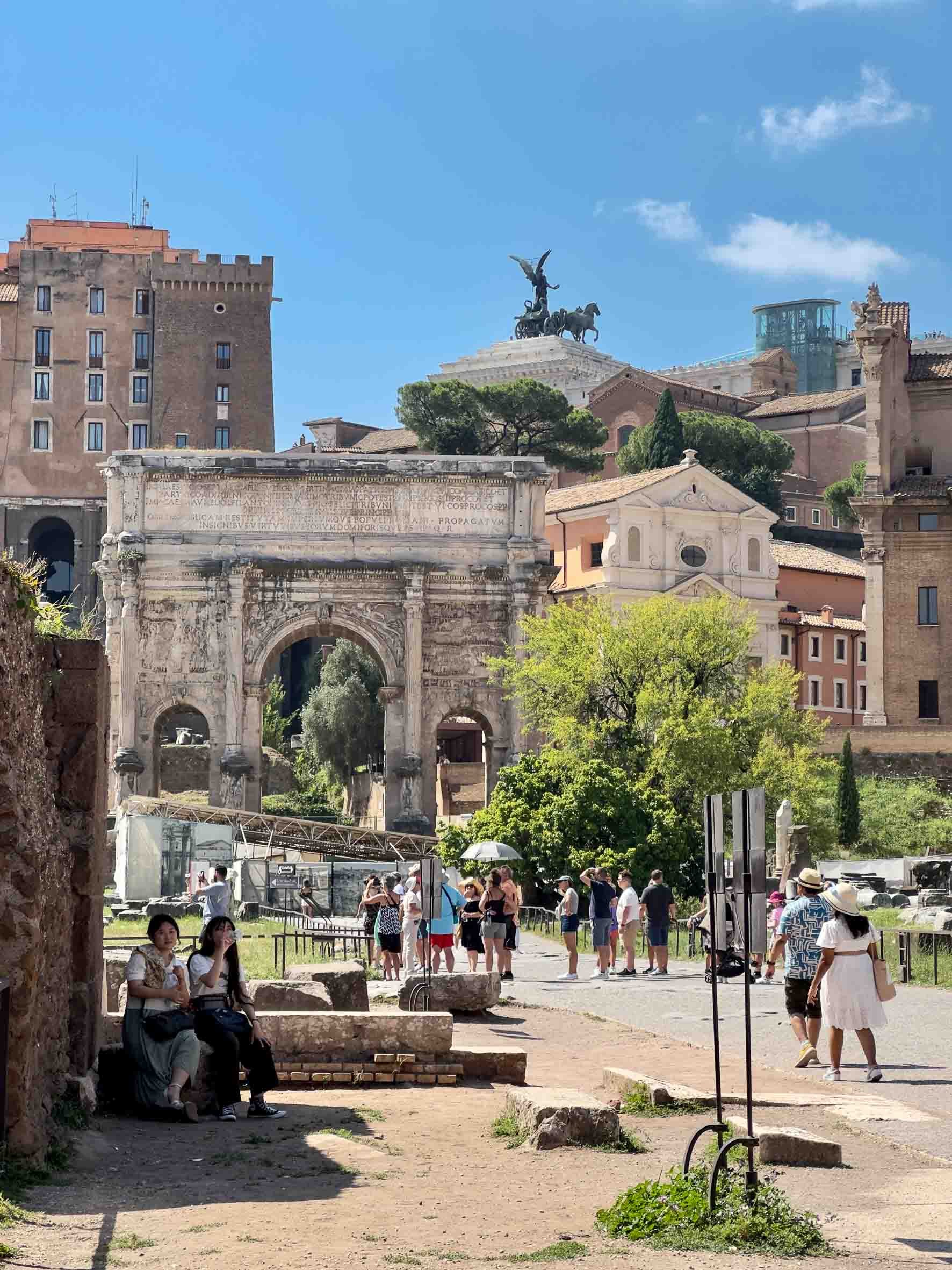 Ruins of an ancient Roman structure with a large arched entrance, surrounded by tourists, with historic buildings and a monument with a chariot and winged figure in the background.