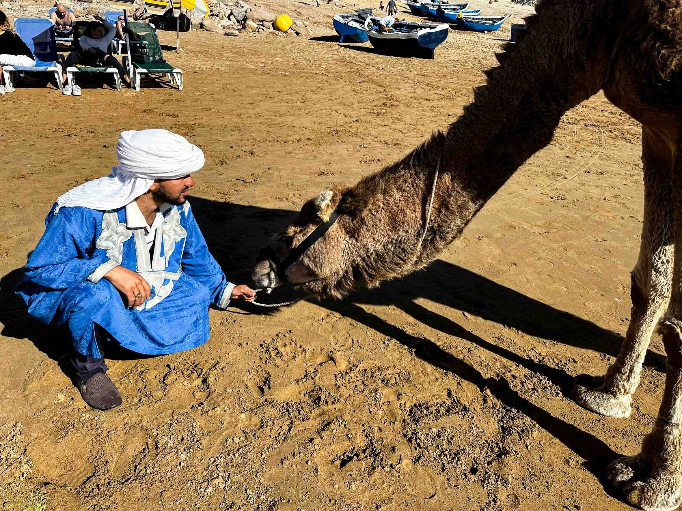 A man dressed in blue traditional clothing and a white head covering is kneeling on sandy ground and holding a camel's bridle, while the camel is extending its head towards the man. In the background, there are beach chairs, people, and small boats o
