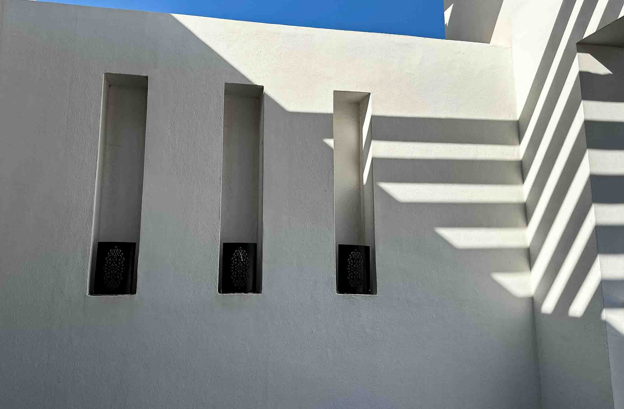 White building with three tall, narrow windows and shadows cast by a staircase on the right side, under a clear blue sky.
