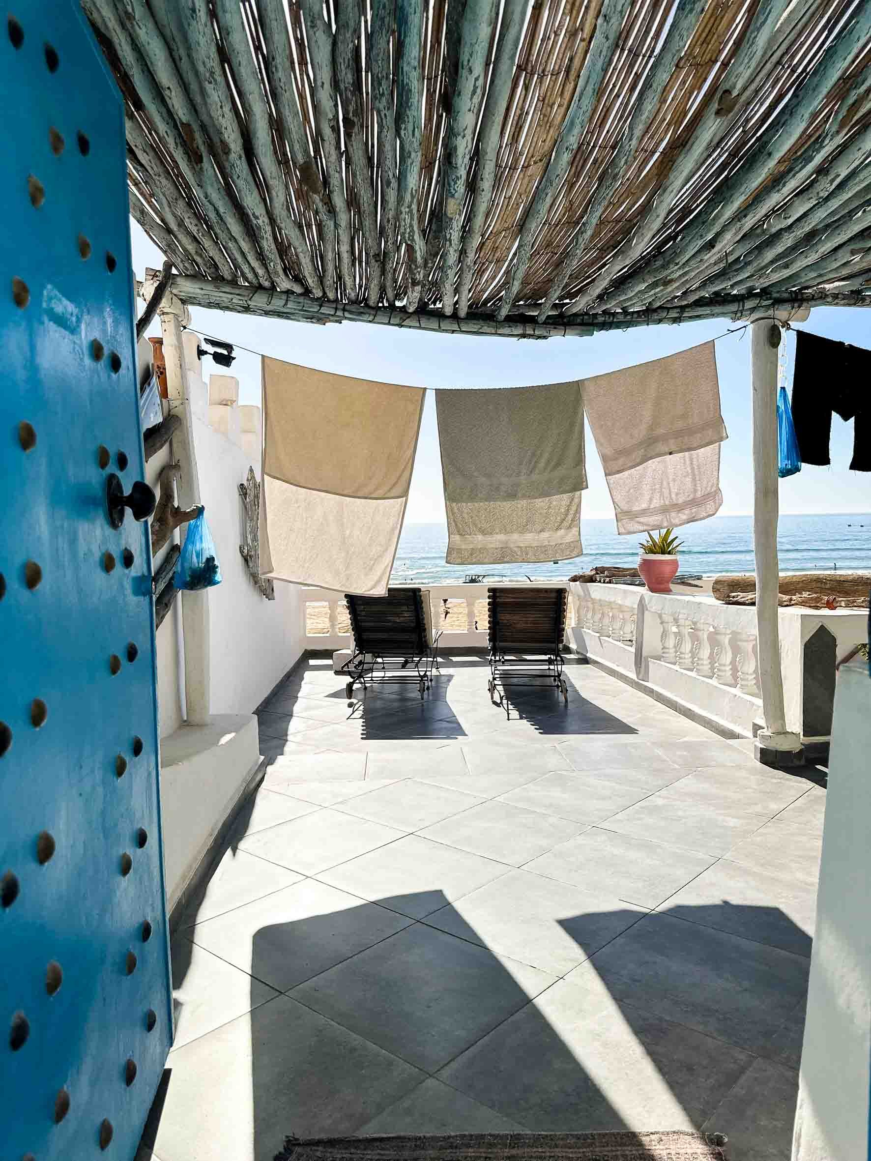 Beachside terrace with two lounge chairs facing the ocean, laundry hanging on a line, potted plant, and shaded by a rustic bamboo roof.