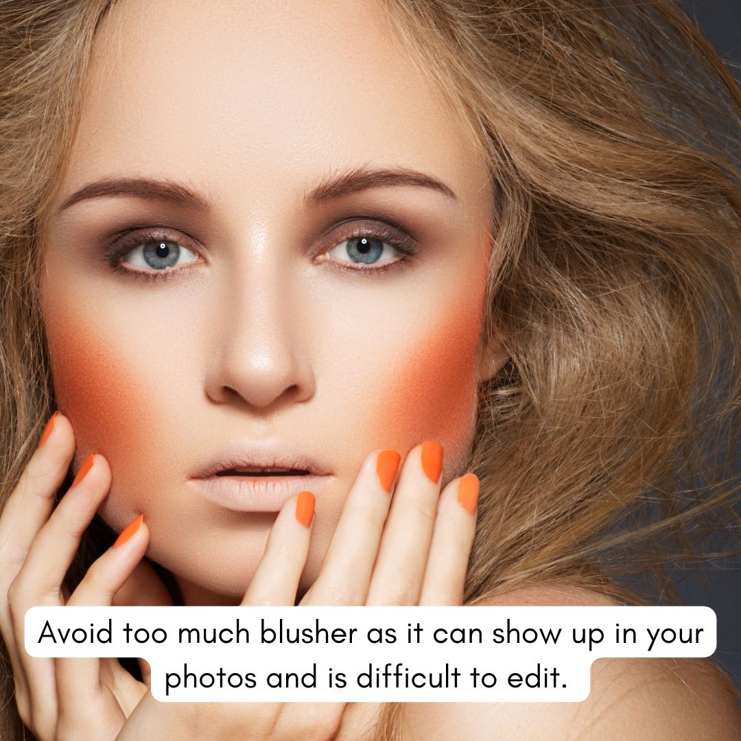 Close-up of a woman with light skin, striking blue eyes, and wavy blonde hair, with orange blush on her cheeks and orange nail polish on her fingers, touching her face.