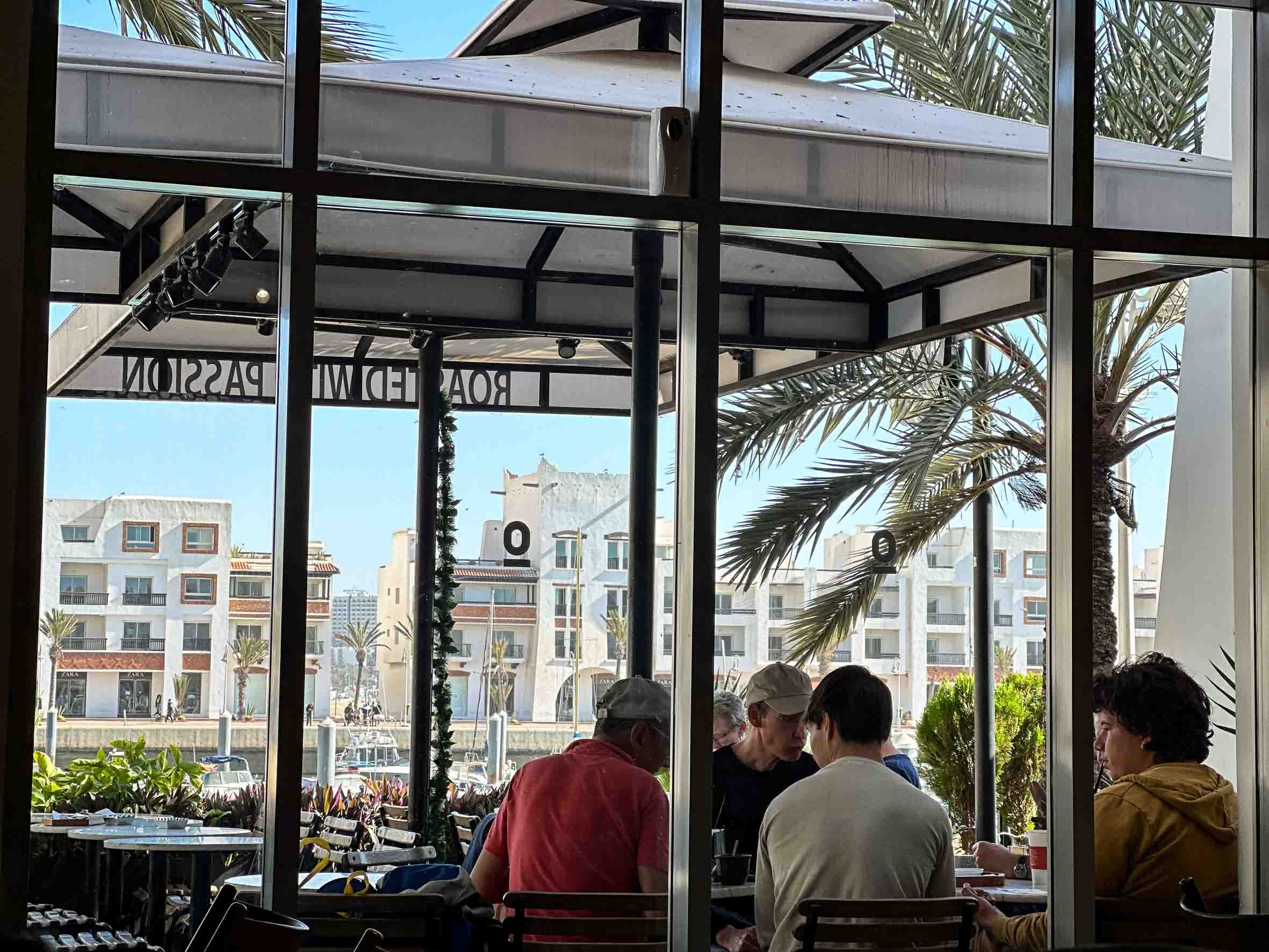 People sitting at a table inside a restaurant with large windows overlooking palm trees and white buildings in a sunny outdoor setting.